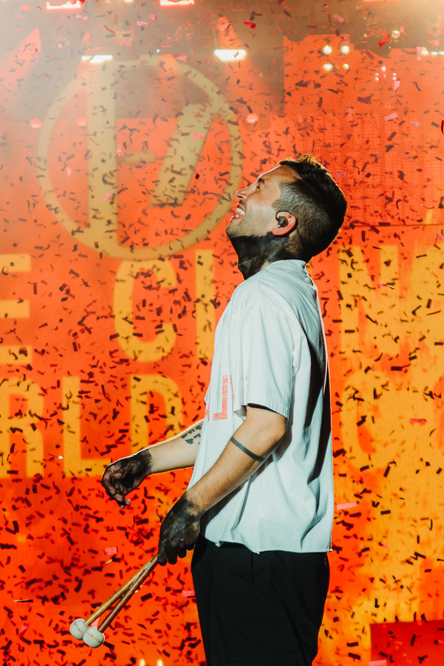 Performer stands on stage against vibrant orange backdrop with dramatic lighting effects during concert performance.
