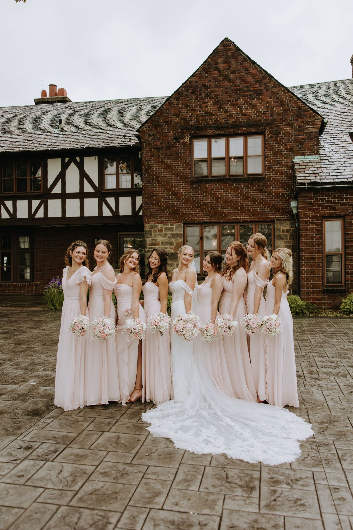 A wedding party in blush pink bridesmaid dresses poses in front of a Tudor-style brick building on a cloudy day.
