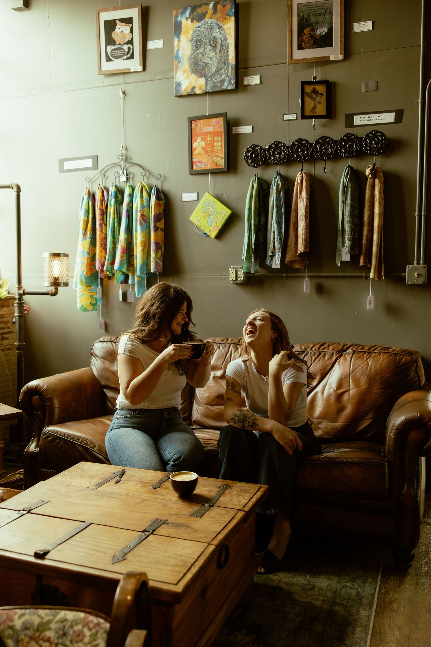Two people relax on a brown leather couch in a cozy room with artwork and clothing displays on the walls.