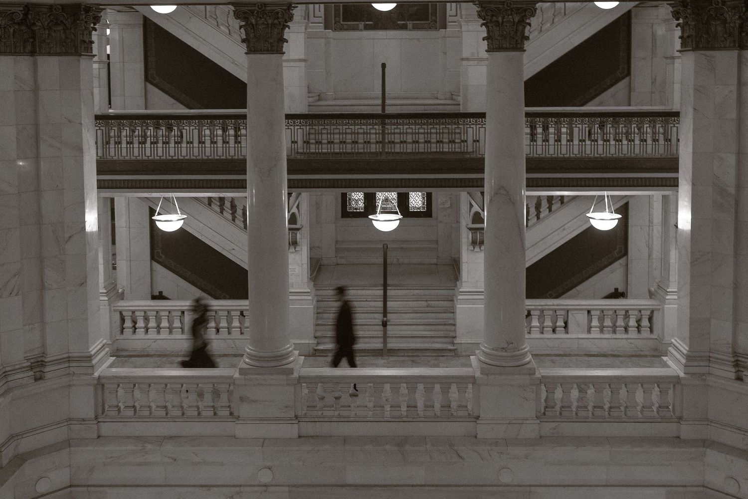 A blurred figure walks through an elegant marble hallway with classical columns and balustrades in black and white.