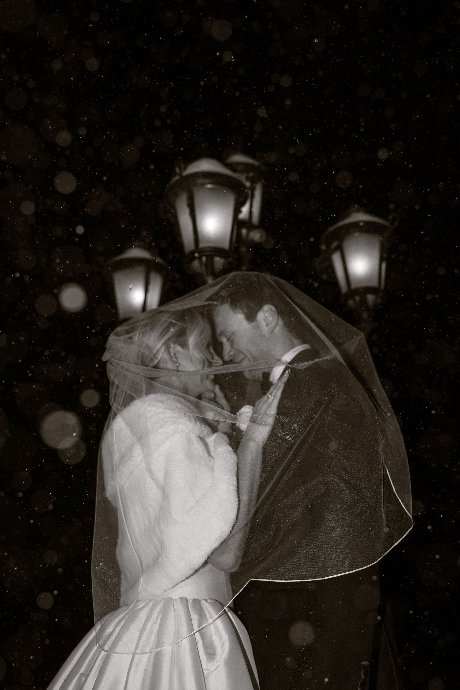 A romantic winter wedding portrait under a lamp post in falling snow.