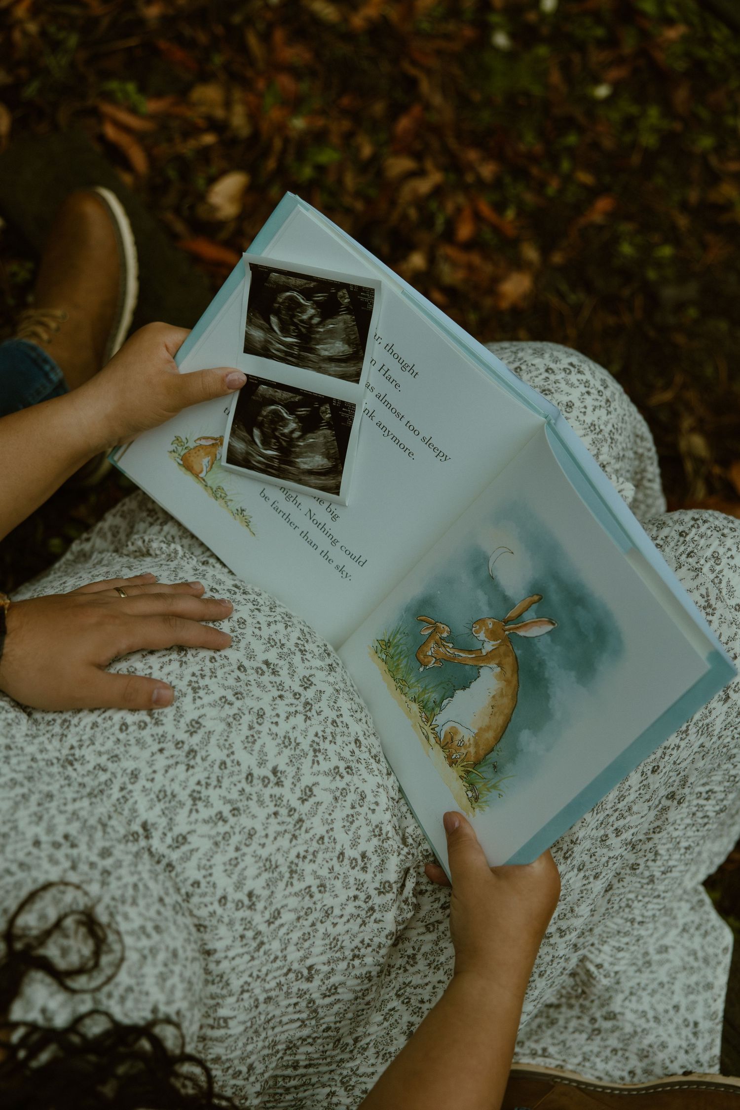 Someone holds a baby book open showing ultrasound images and illustrations while sitting on a patterned blanket outdoors.