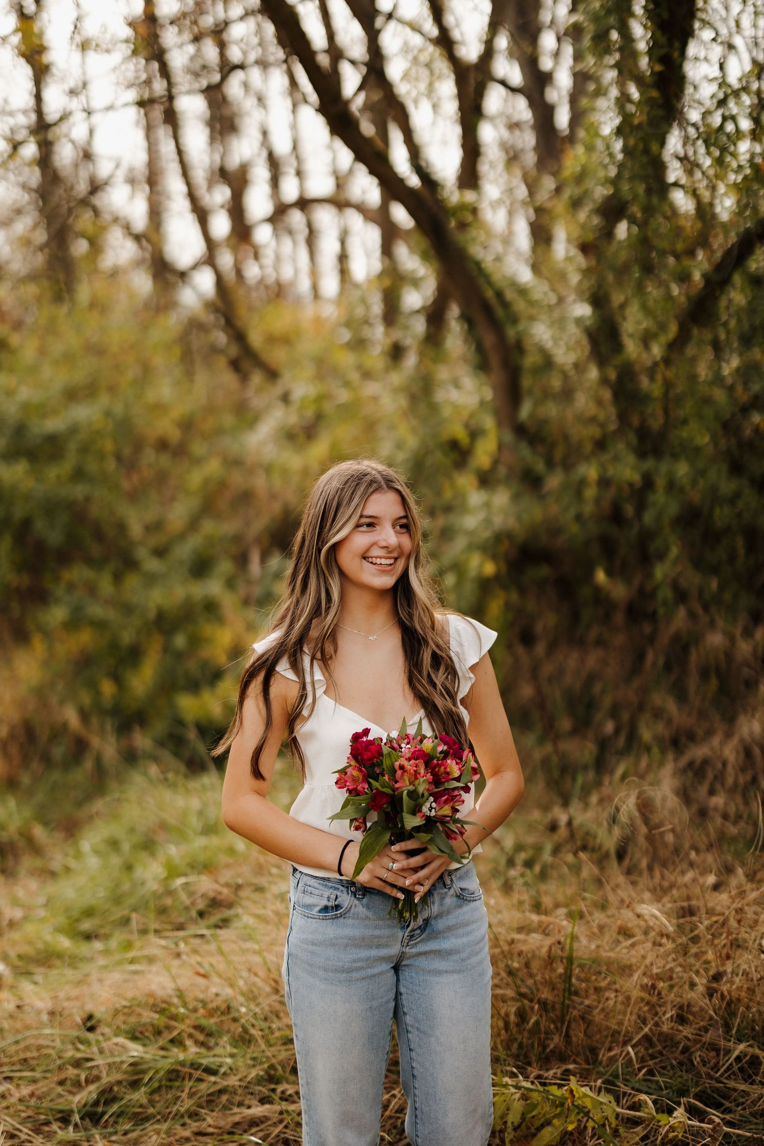 A woman in casual attire holding a vibrant red bouquet poses in a wooded autumn setting.