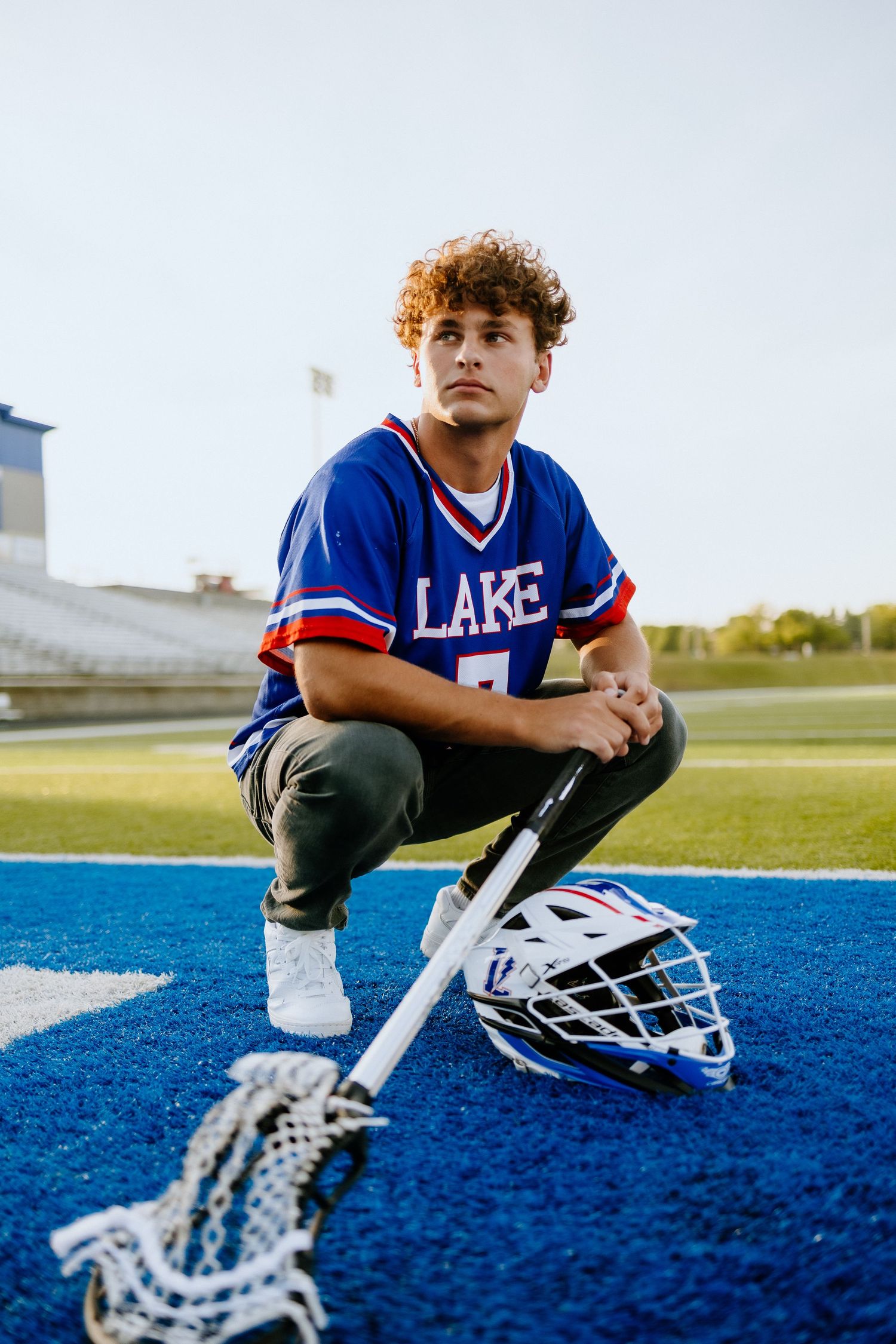 Lacrosse player in blue uniform kneels on field with helmet and stick.