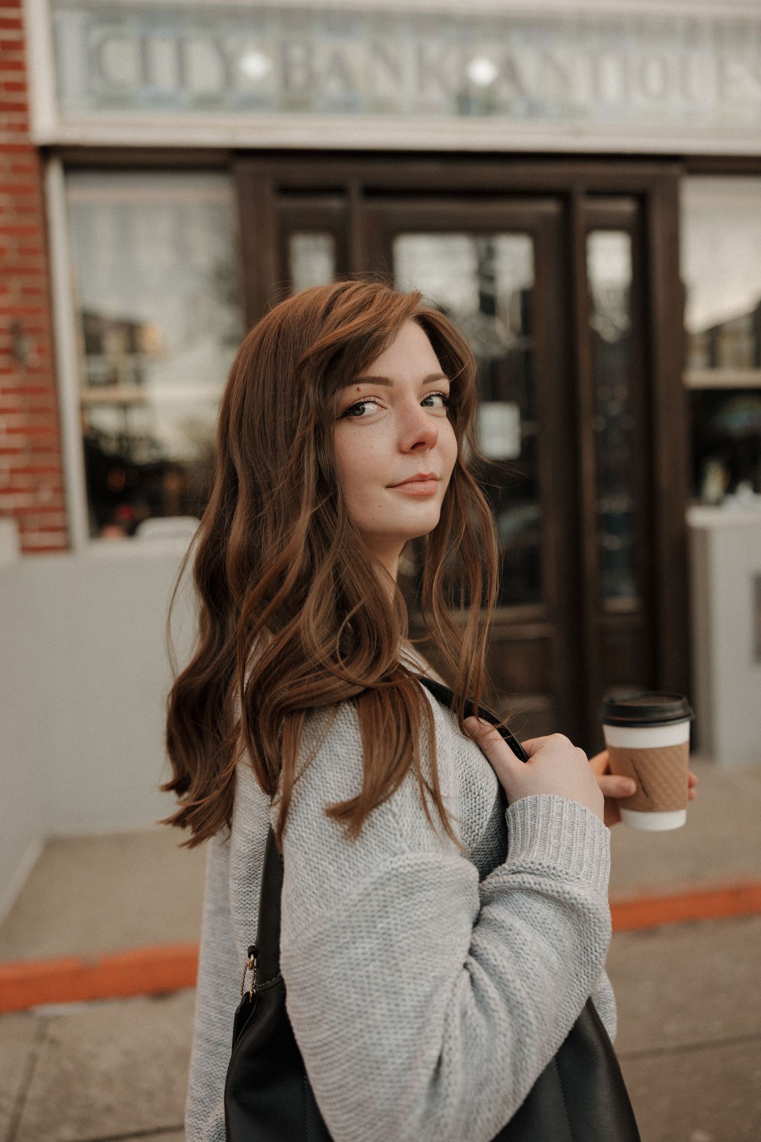 Person in gray sweater holding coffee cup outside storefront.