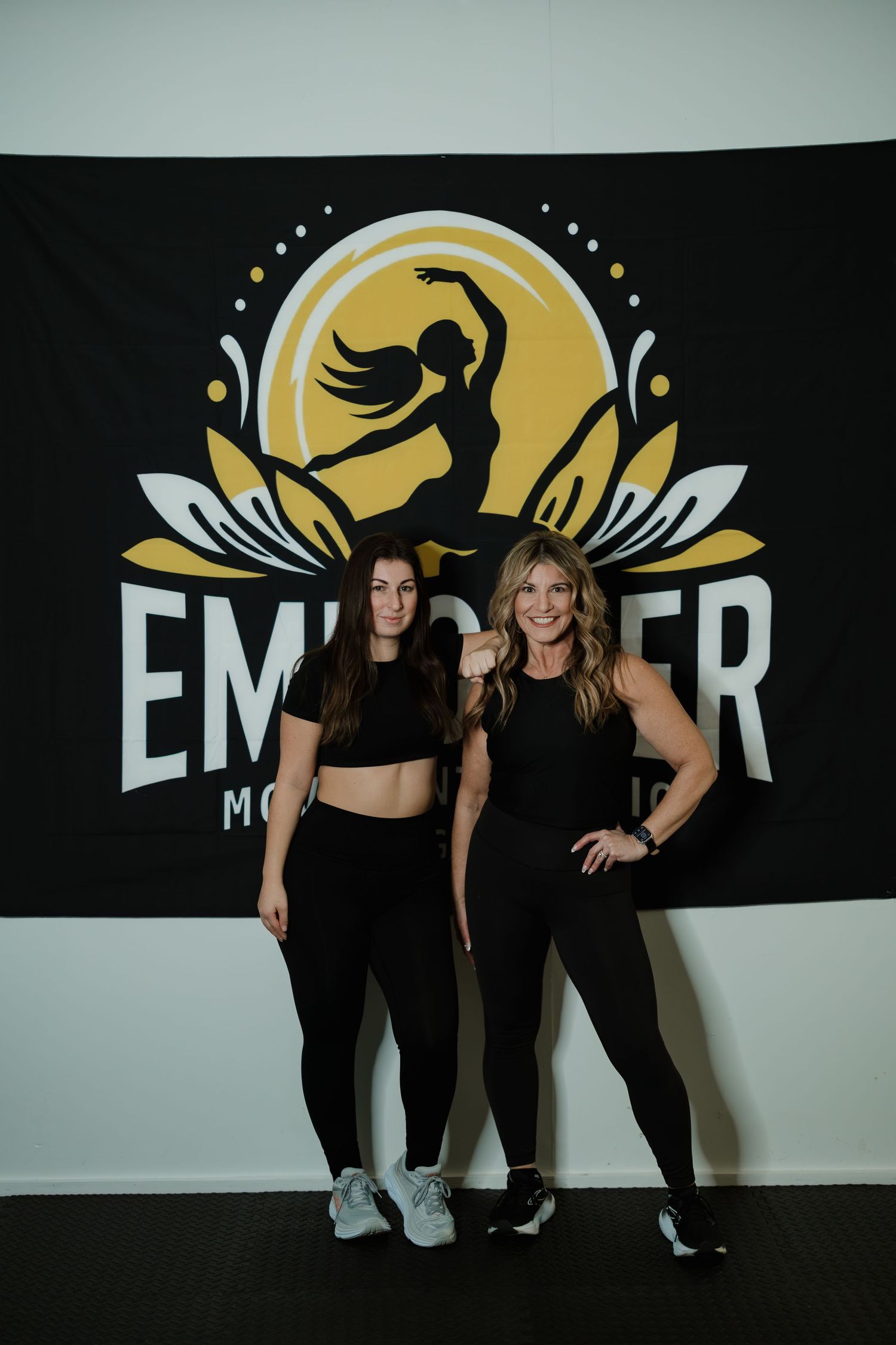 Two people posing in front of a black banner with gold dance studio logo.