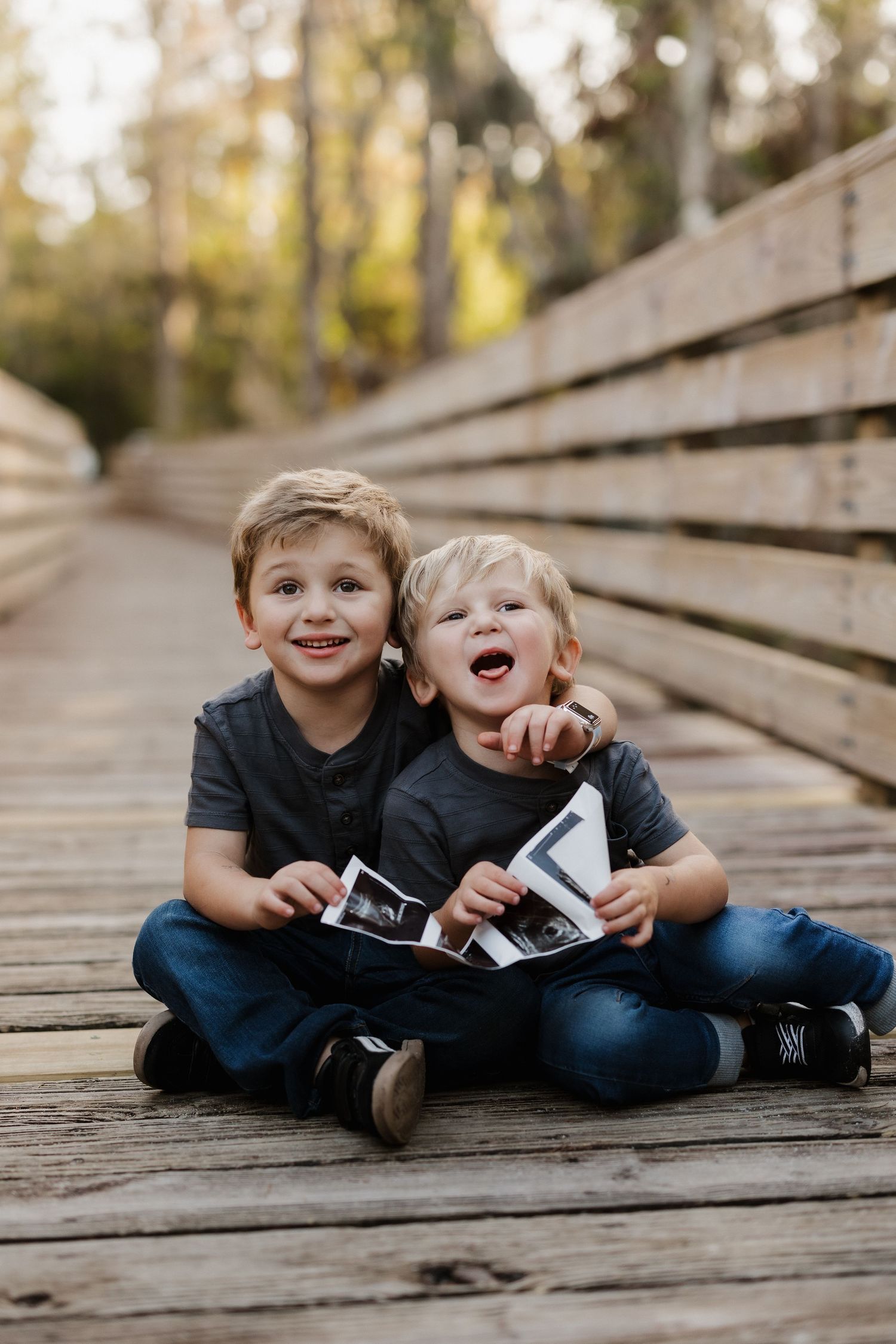 Two young children sit on a wooden boardwalk sharing photos and laughing together.