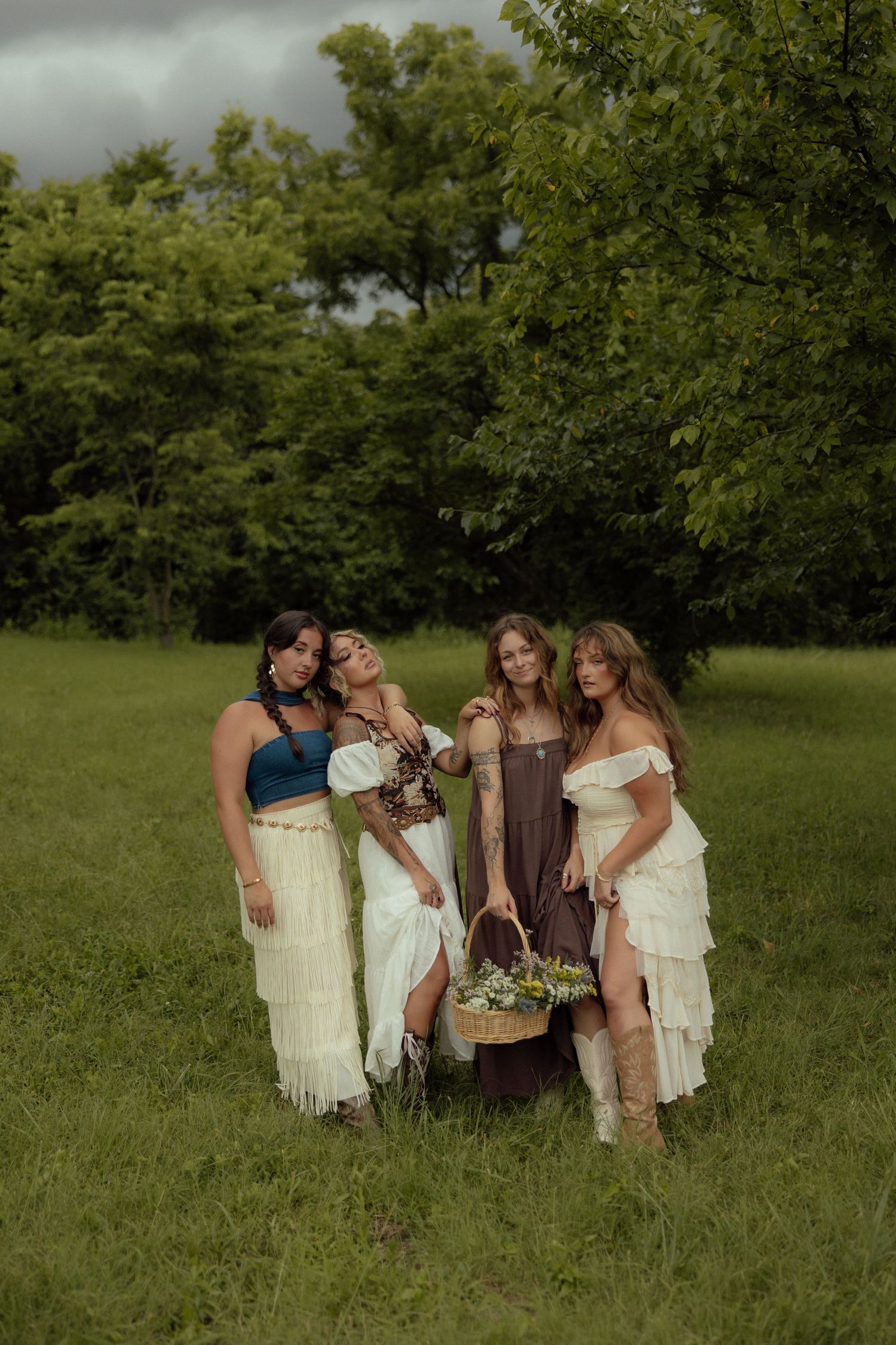 Four women in bohemian-style dresses pose together in a lush green field holding floral bouquets and baskets.