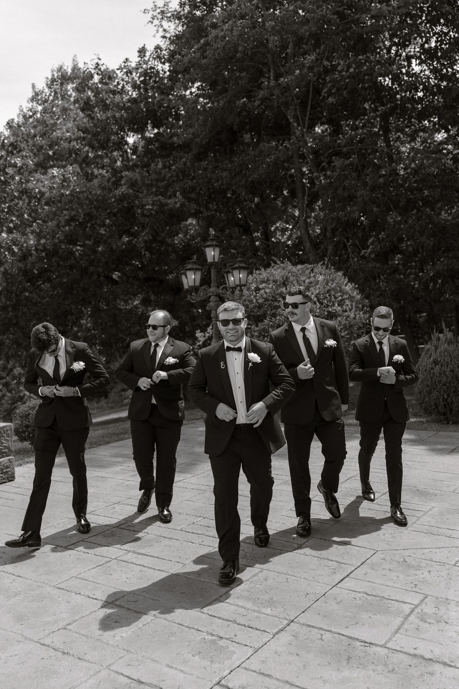 A group in formal black suits walking together in a line on a stone pathway surrounded by trees.