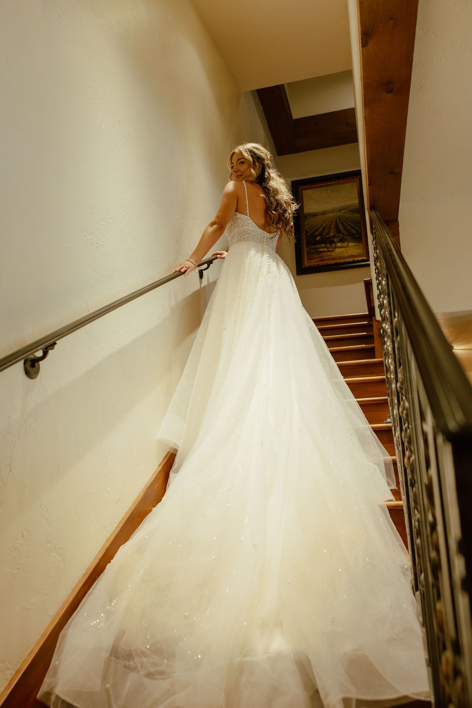 A bride in a flowing white wedding gown descends an elegant staircase while holding the railing.