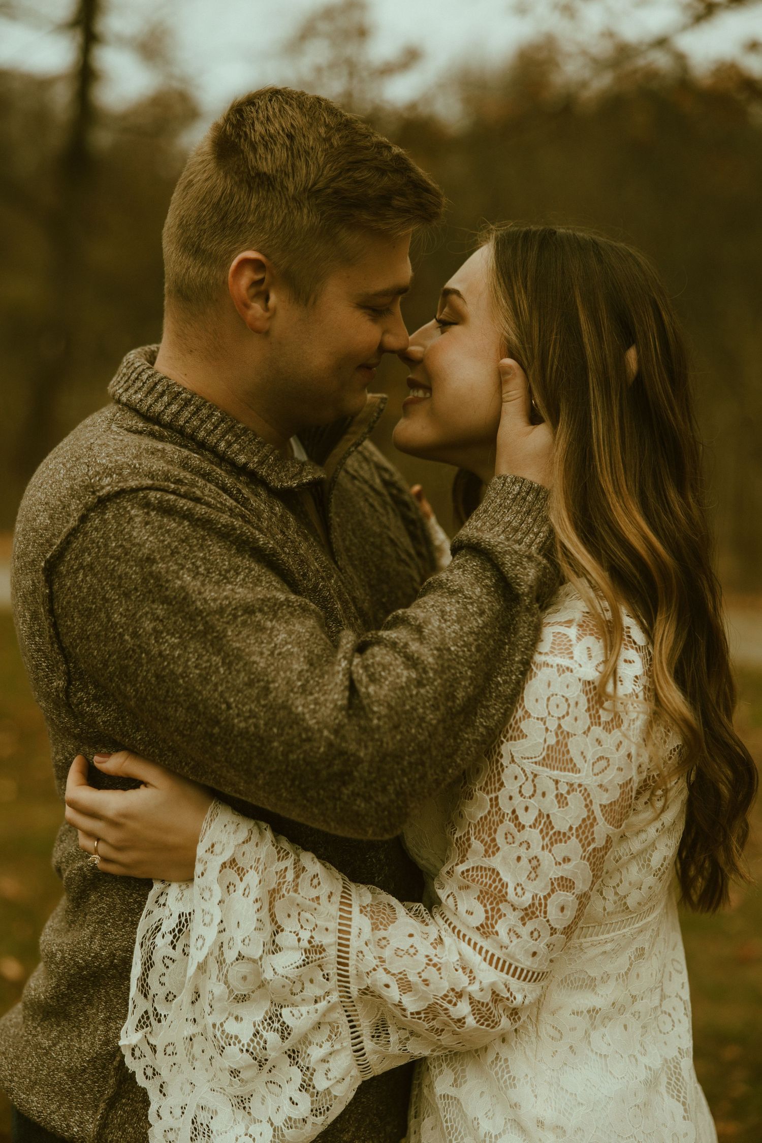 A romantic couple embraces in a golden sunset, with the woman wearing a lace dress and man in a brown sweater.
