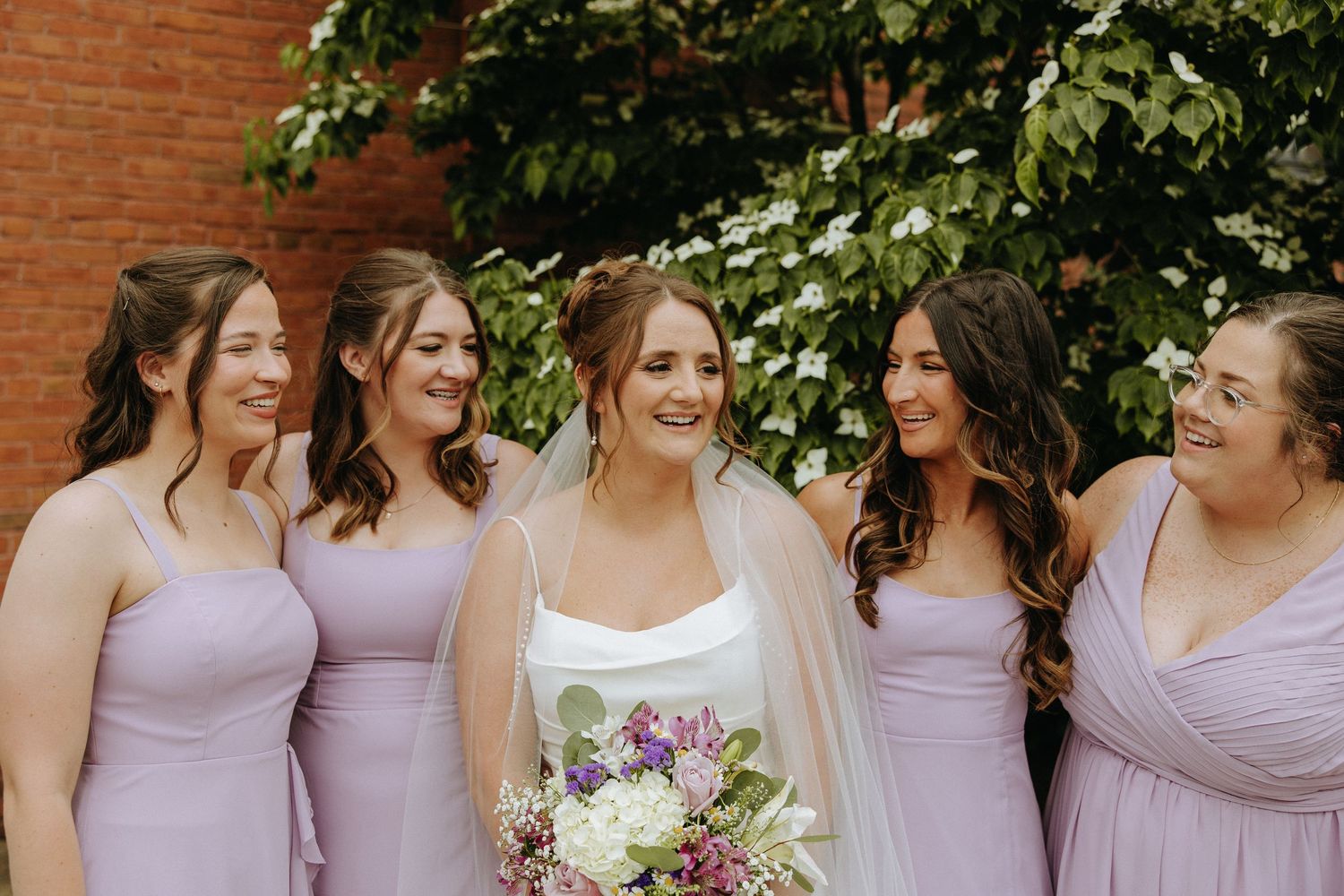 Bridesmaids in lavender dresses gather together outdoors for a wedding photo.