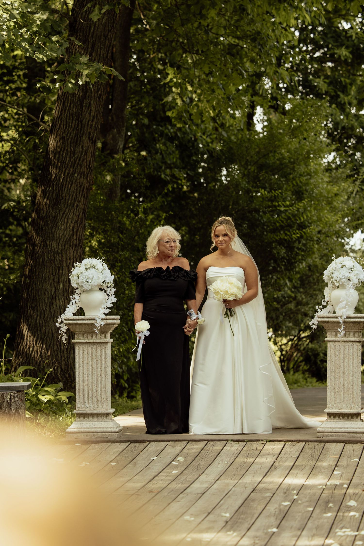 Wedding party poses on an outdoor wooden deck with decorative white columns and lush greenery in background.