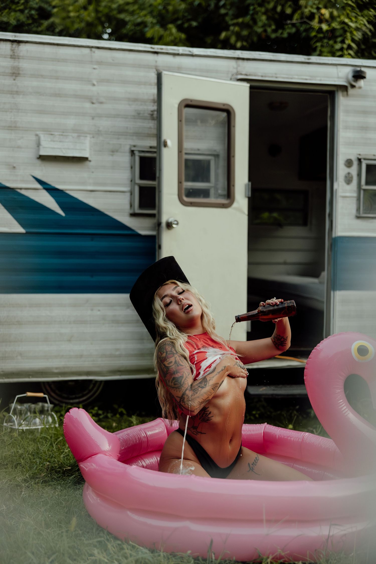 A person lounges and laughs in a pink inflatable pool next to a white trailer on a summer day.