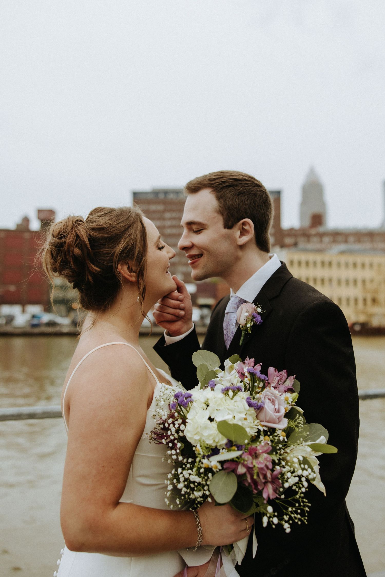 Wedding couple shares romantic moment with floral bouquet against urban waterfront backdrop.