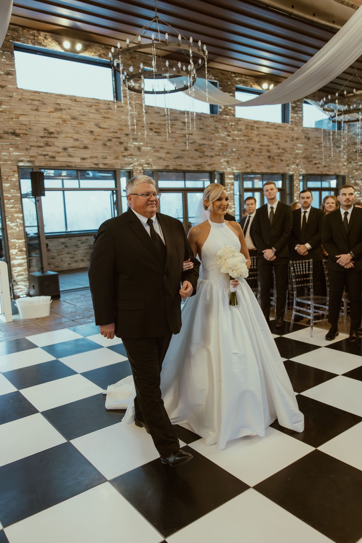 Wedding ceremony on black and white checkered floor with stone wall backdrop and natural lighting.