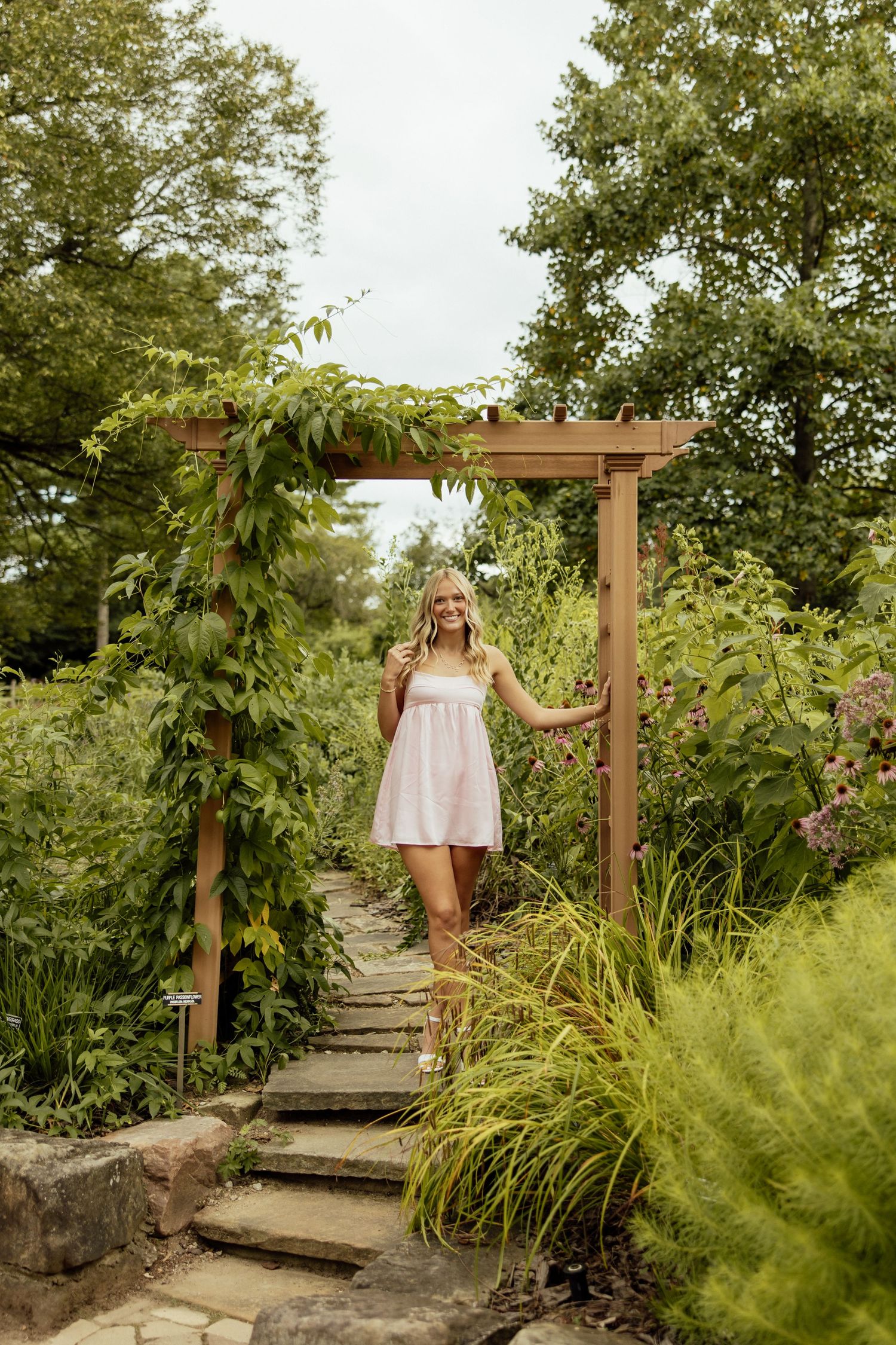 A wooden pathway through a lush garden leads to a rustic pergola surrounded by greenery and ornamental grasses.