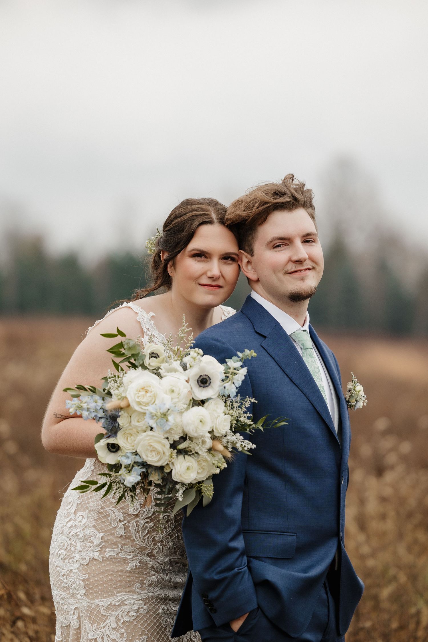 Newlyweds pose in field with white floral bouquet and classic wedding attire.