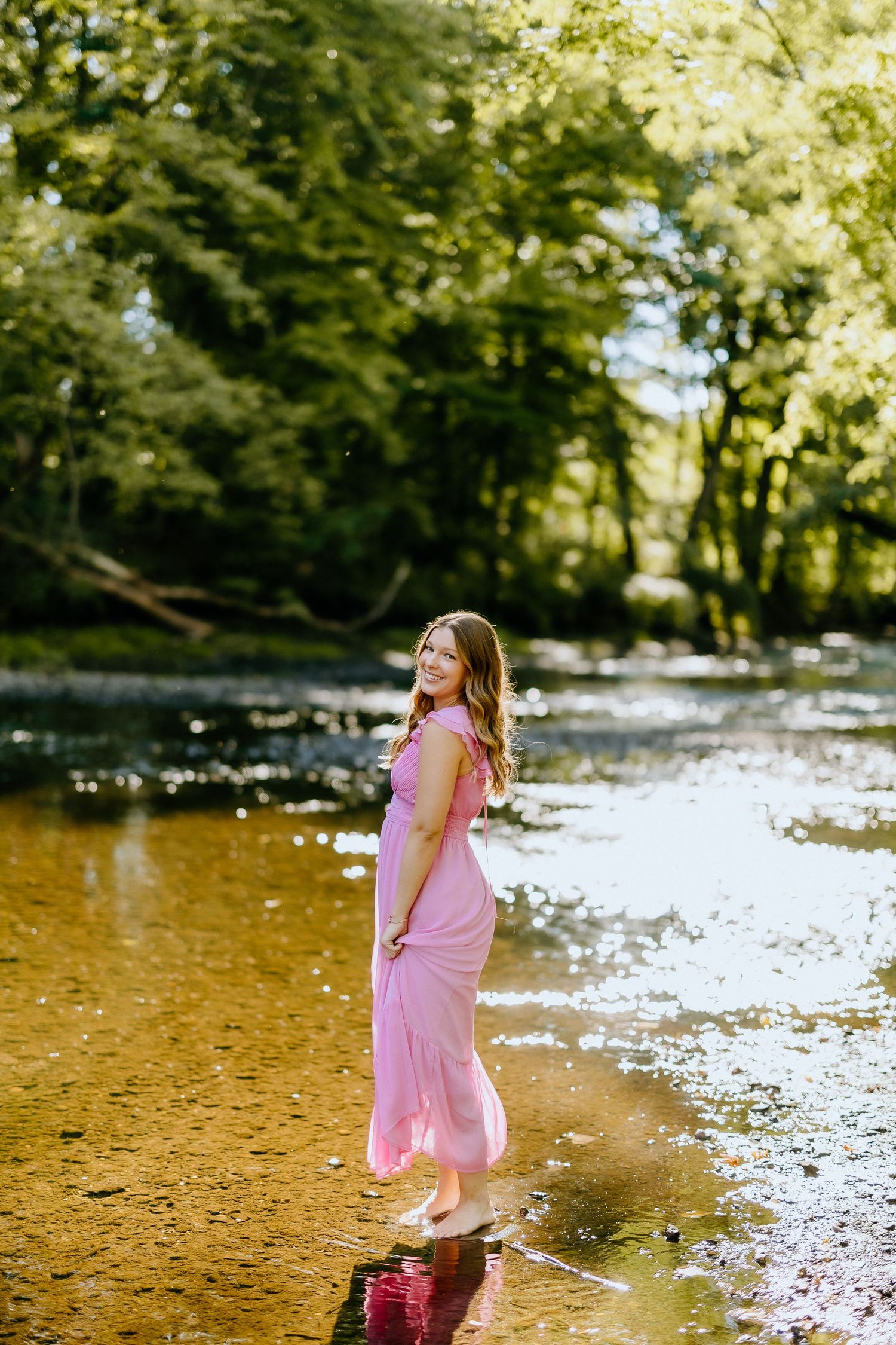 A pink dress catches golden light while standing at the edge of a peaceful stream.