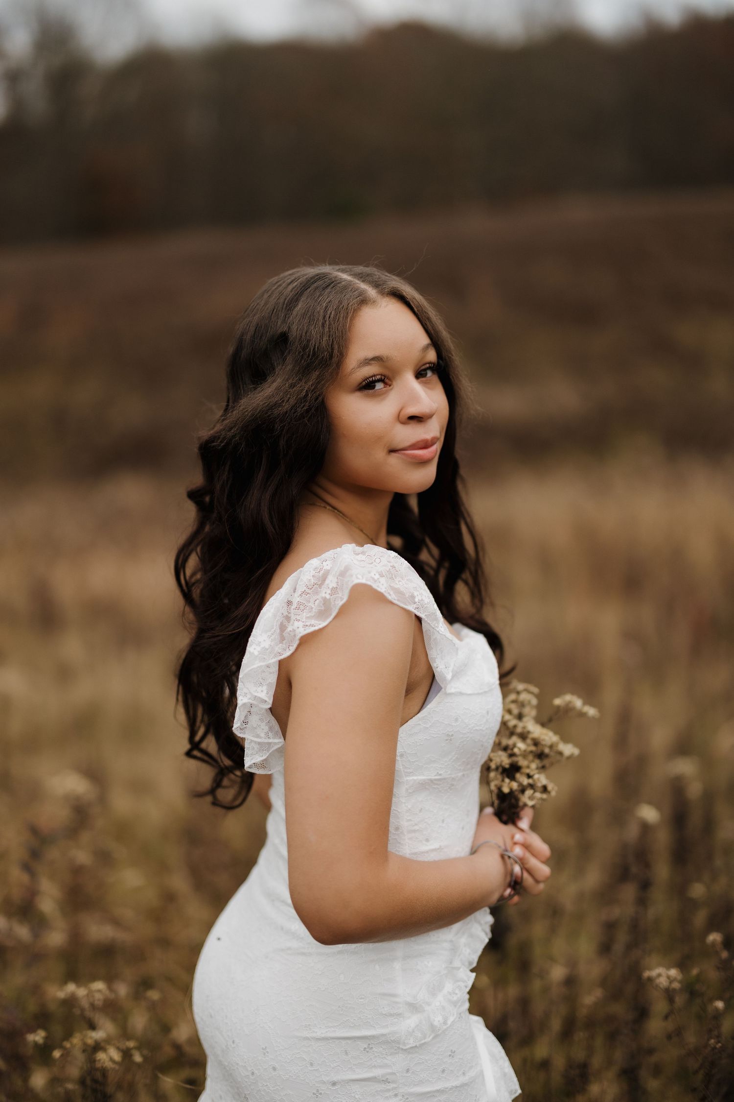 A romantic outdoor portrait shows someone in a lace white dress posing in a golden field at sunset.