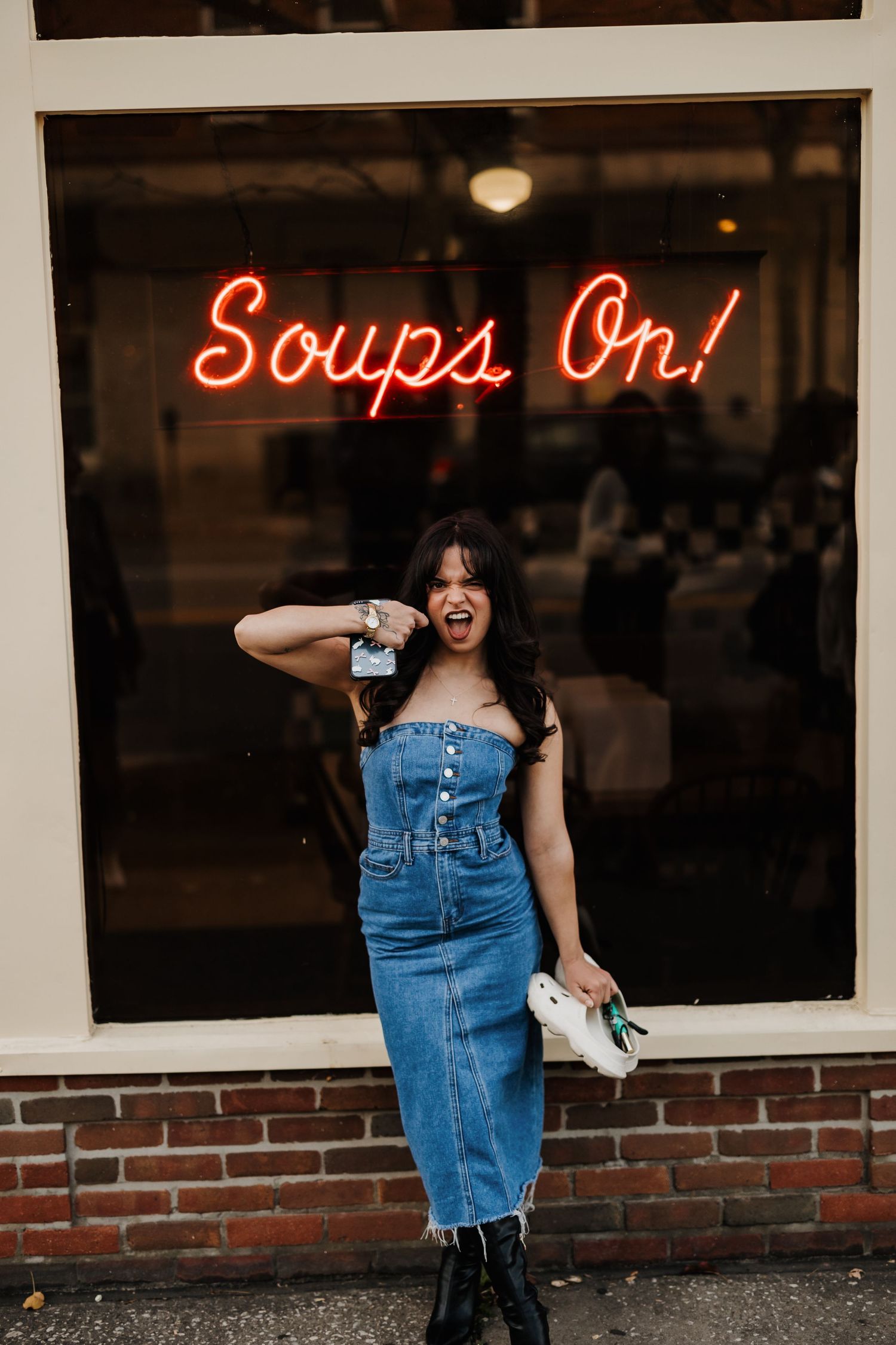 Figure posing in denim dress beneath red neon 'Soups On!' sign at dusk.