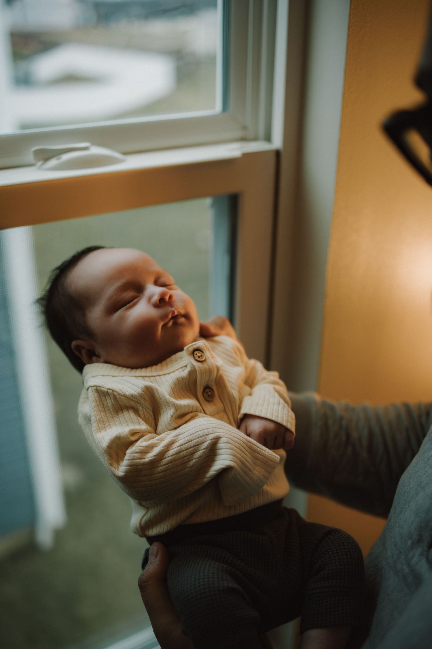 A content baby in a cream sweater relaxes by a window in warm lighting.