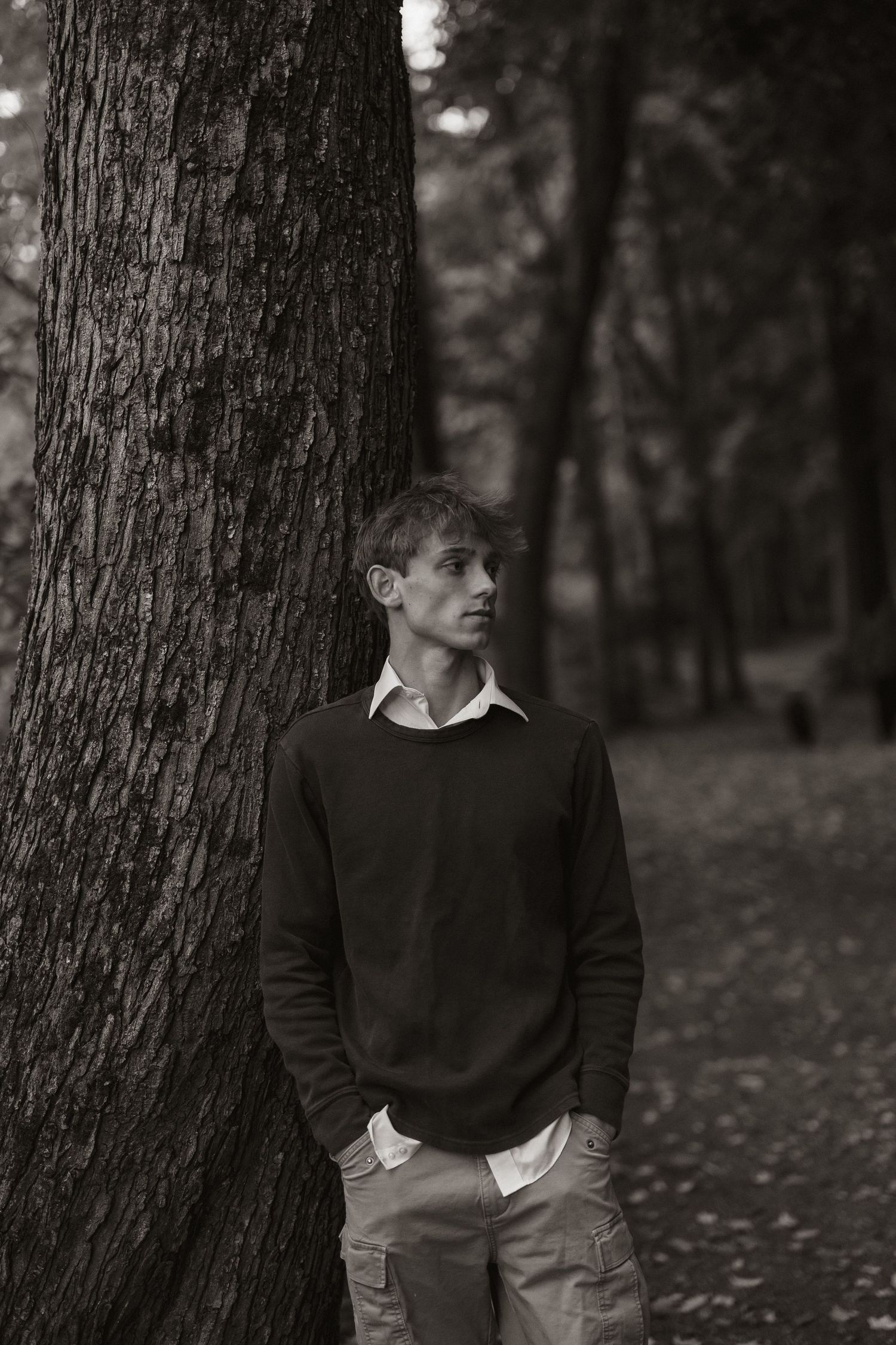 Moody black and white portrait against a large tree trunk in a wooded setting.