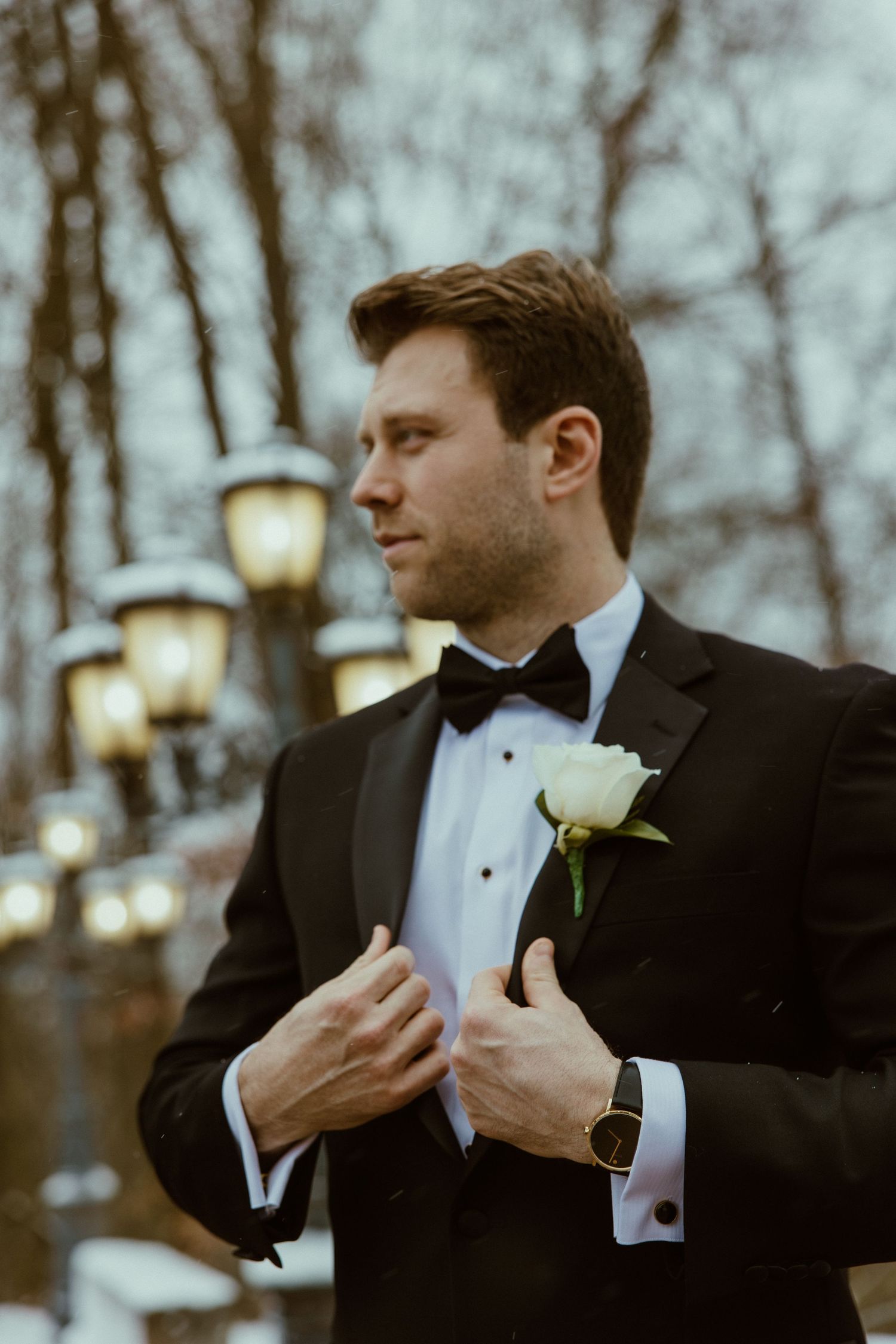 A groom in a black tuxedo adjusts his white boutonniere outdoors.
