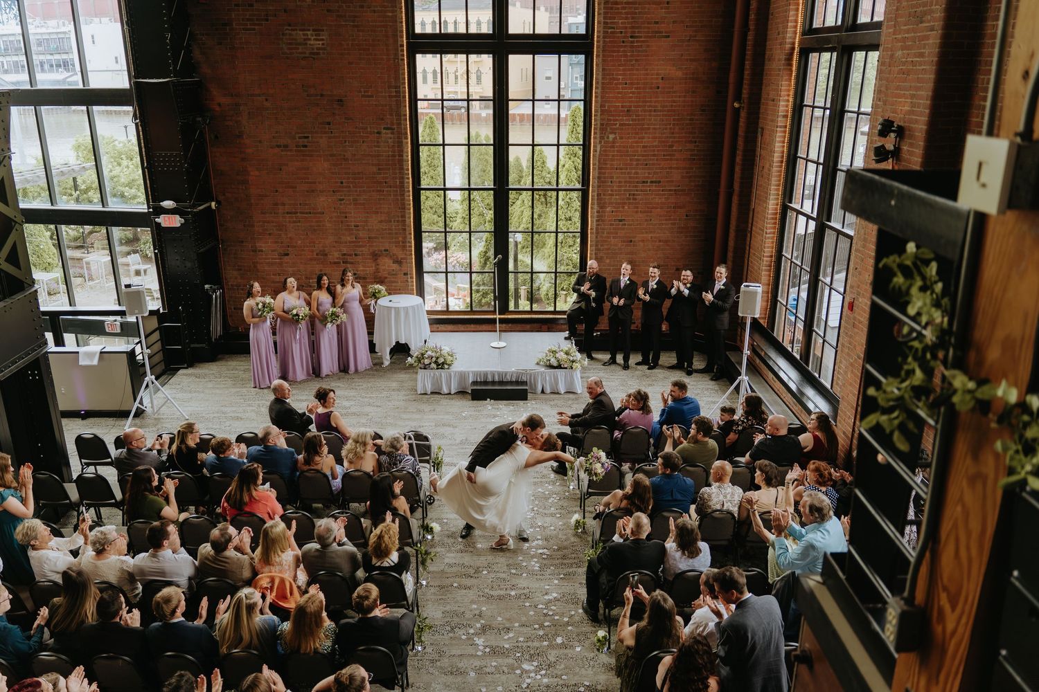 Wide angle view of a wedding ceremony in a rustic brick venue with large windows and gathered guests.