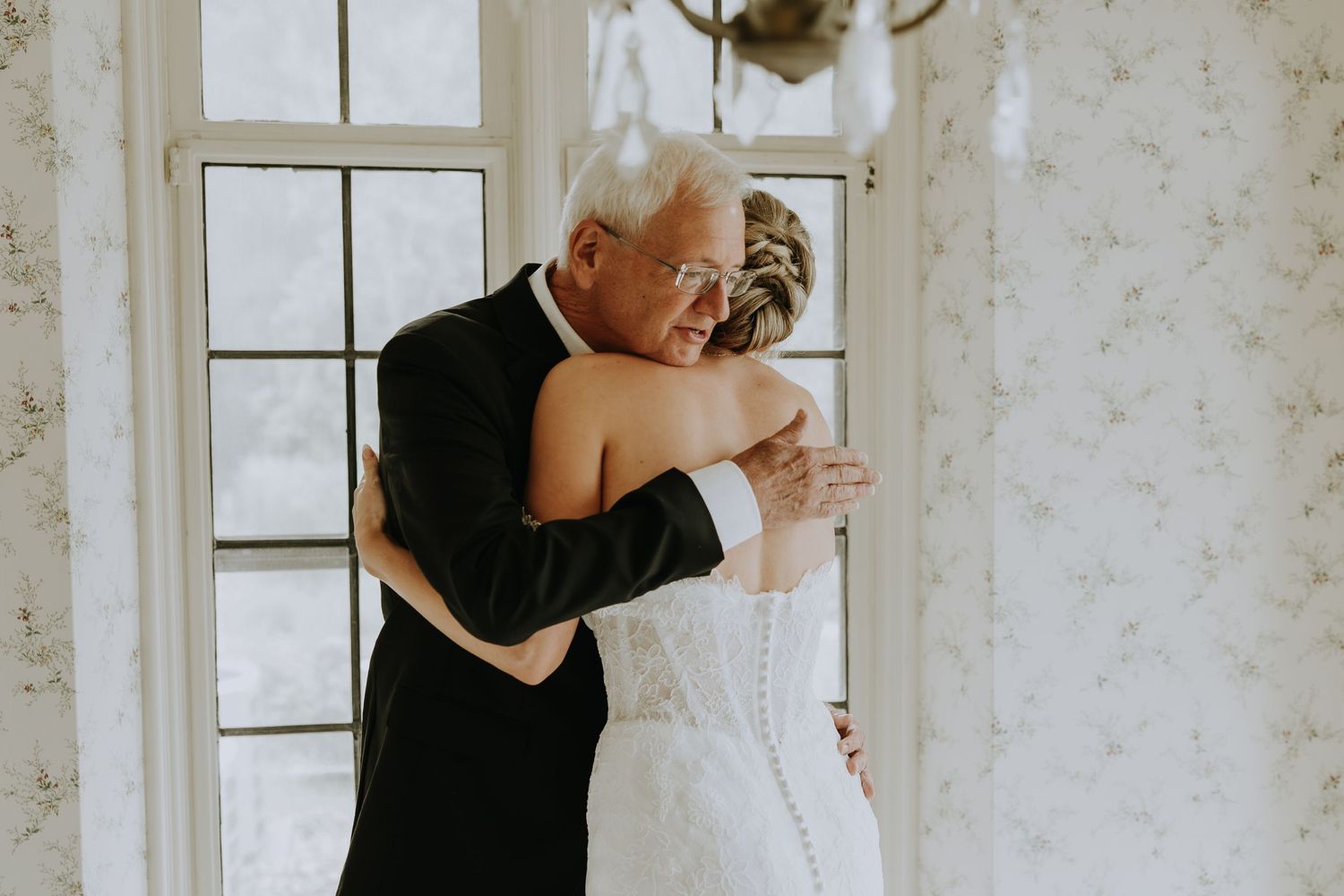 A wedding party shares an emotional moment together in a sun-lit room with floral wallpaper.