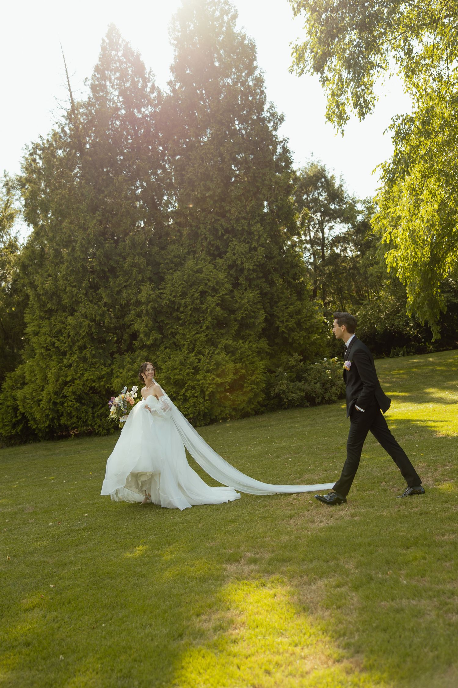 A couple walking on a sunlit lawn with a flowing white wedding dress train trailing behind them.