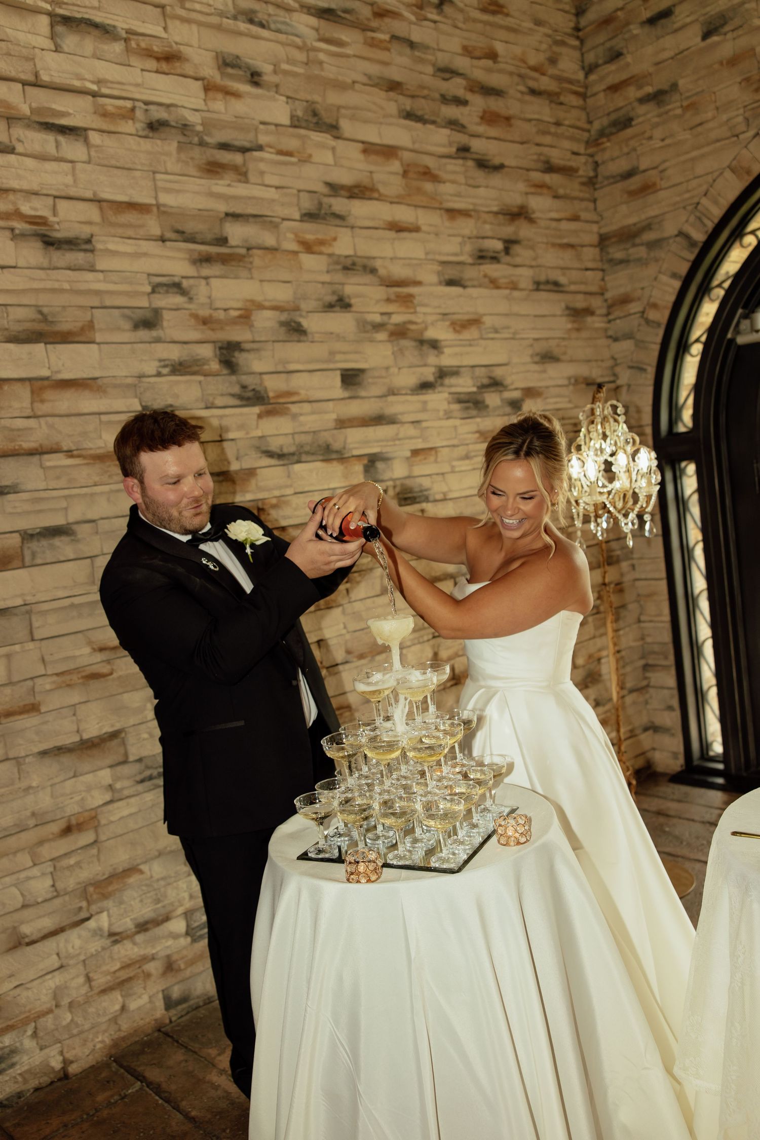 A couple builds a champagne tower by pouring bubbly into stacked glasses at their wedding reception.