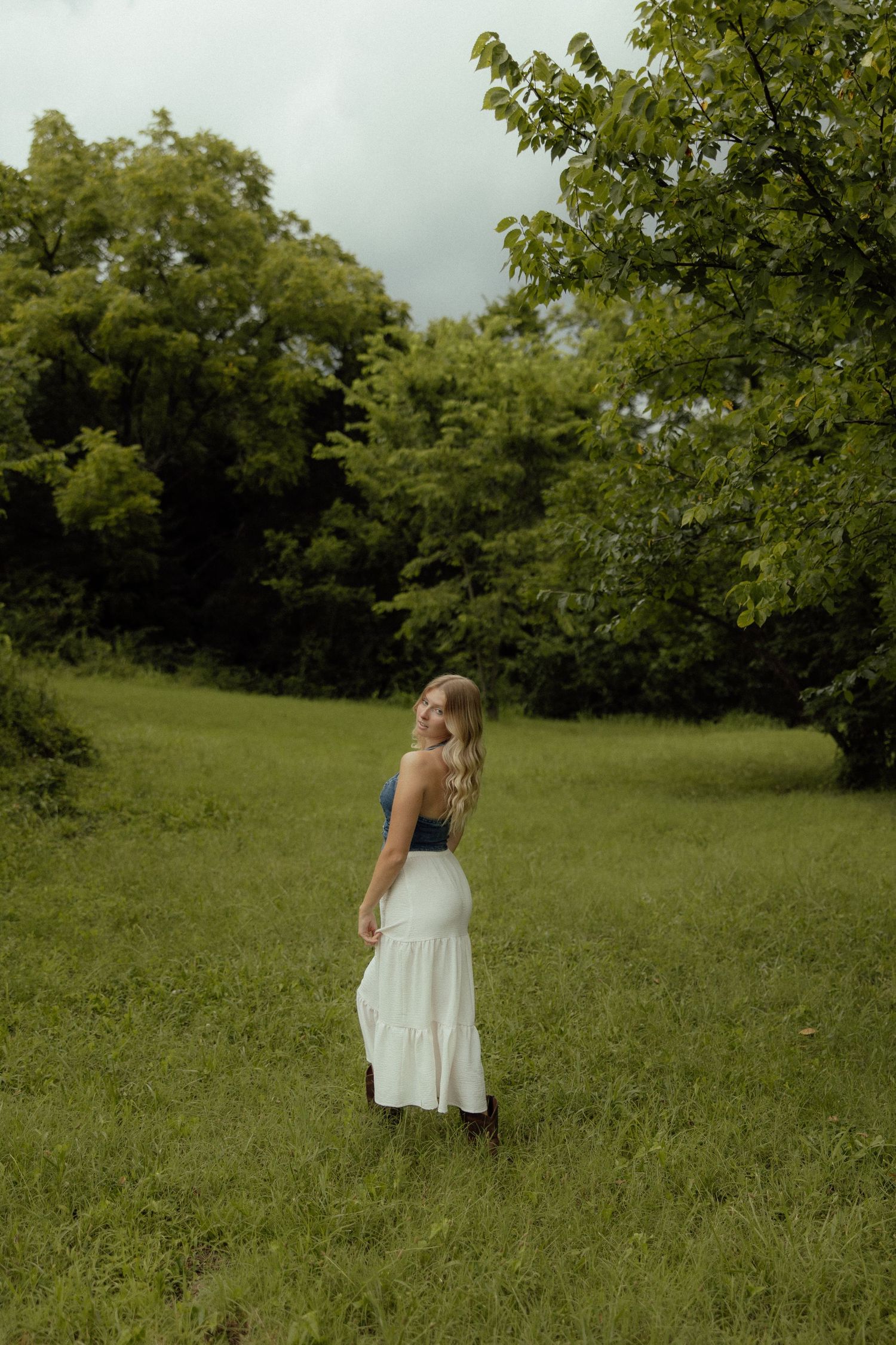 A person in a white sundress twirls playfully in a lush green meadow surrounded by trees on an overcast day.