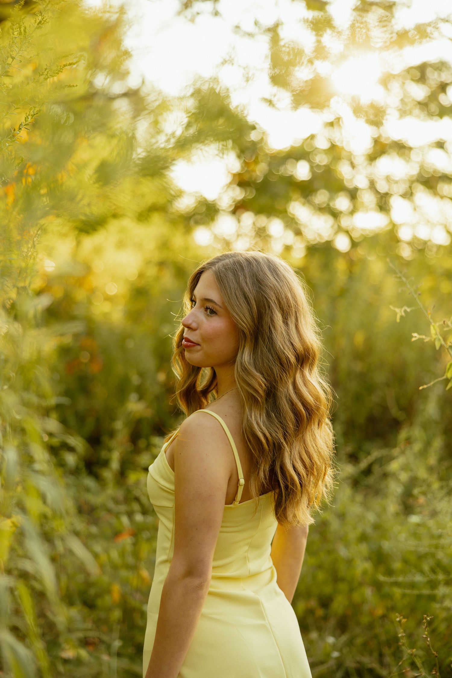 A person in a yellow dress poses against a dreamy golden sunset background in a natural outdoor setting.