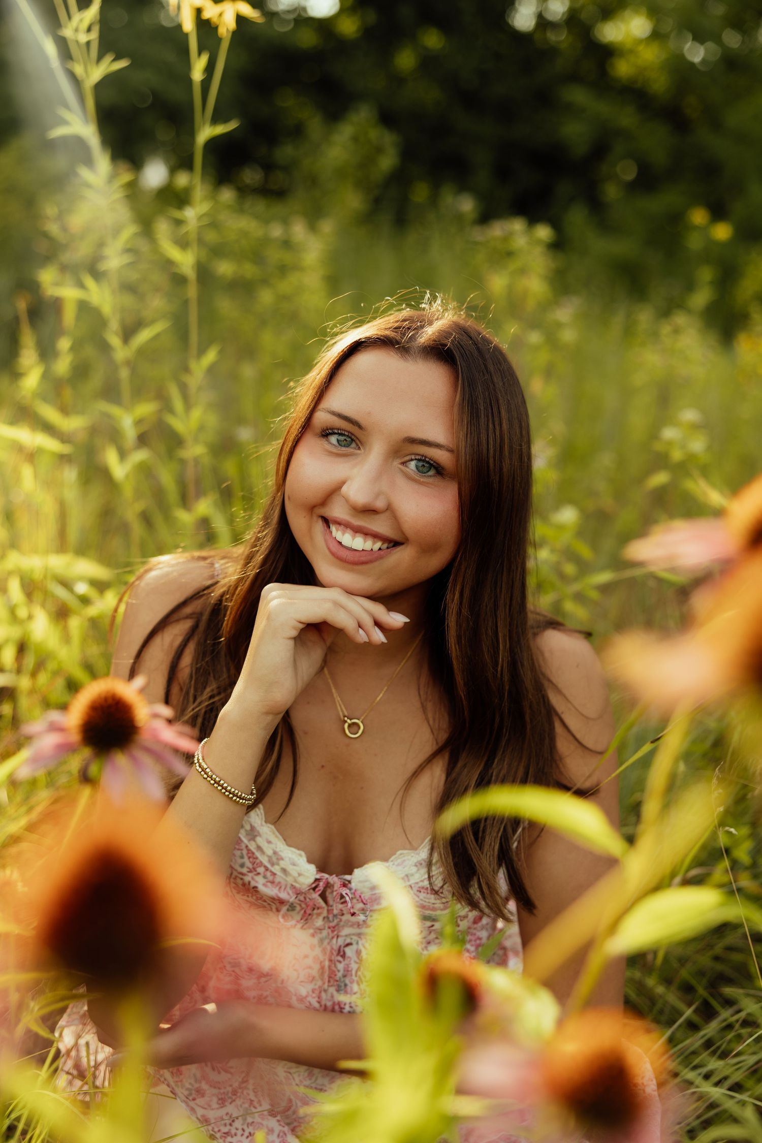 A person poses in a floral dress among tall wildflowers and greenery in a summer meadow setting.