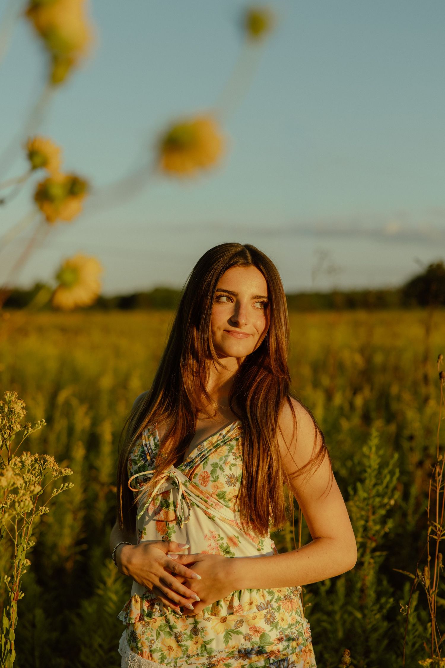 A person in a floral dress stands thoughtfully in a golden field at sunset with wildflowers swaying in the breeze.