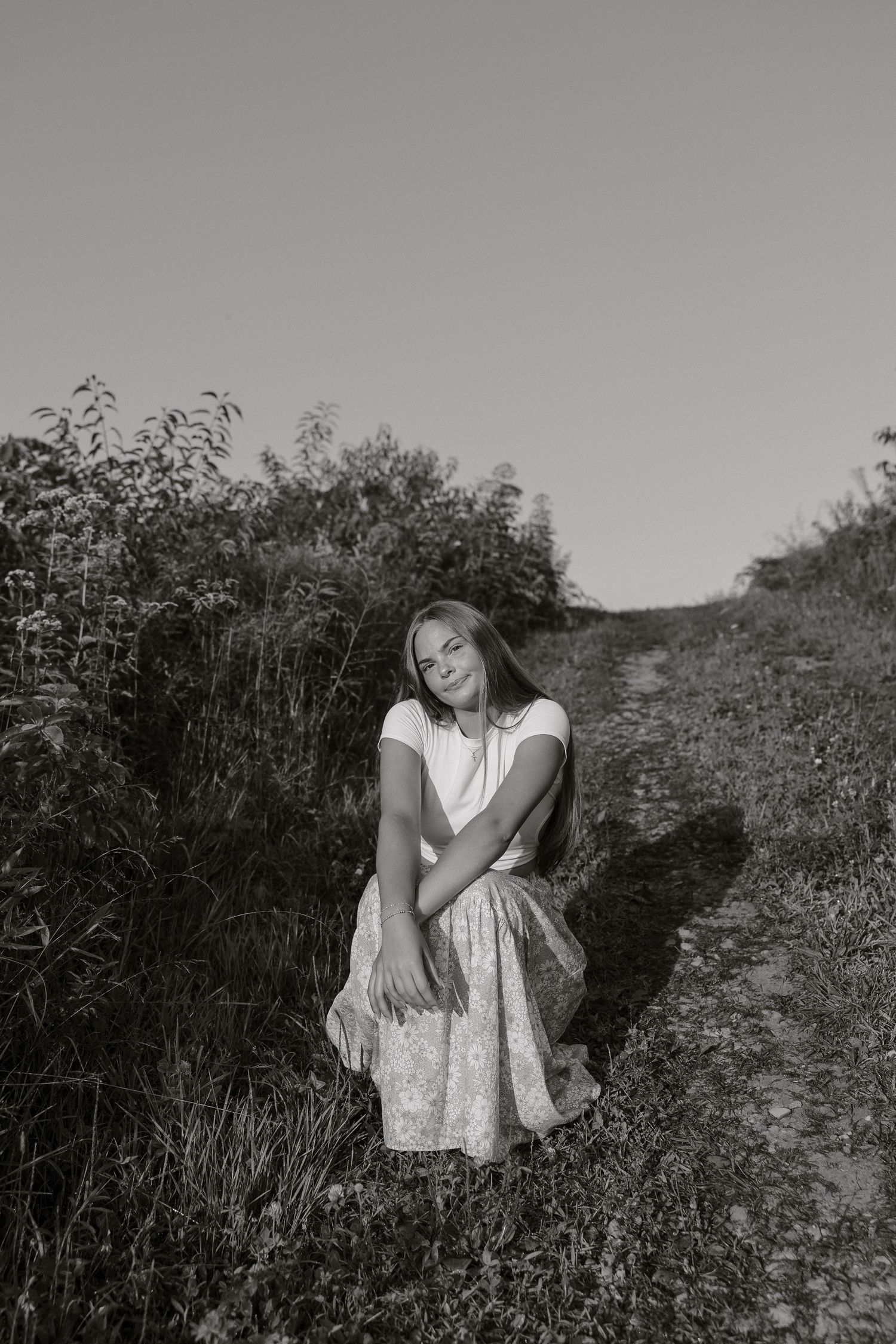 A series of black and white photographs showing a person in a light dress sitting among wildflowers on a hillside path.