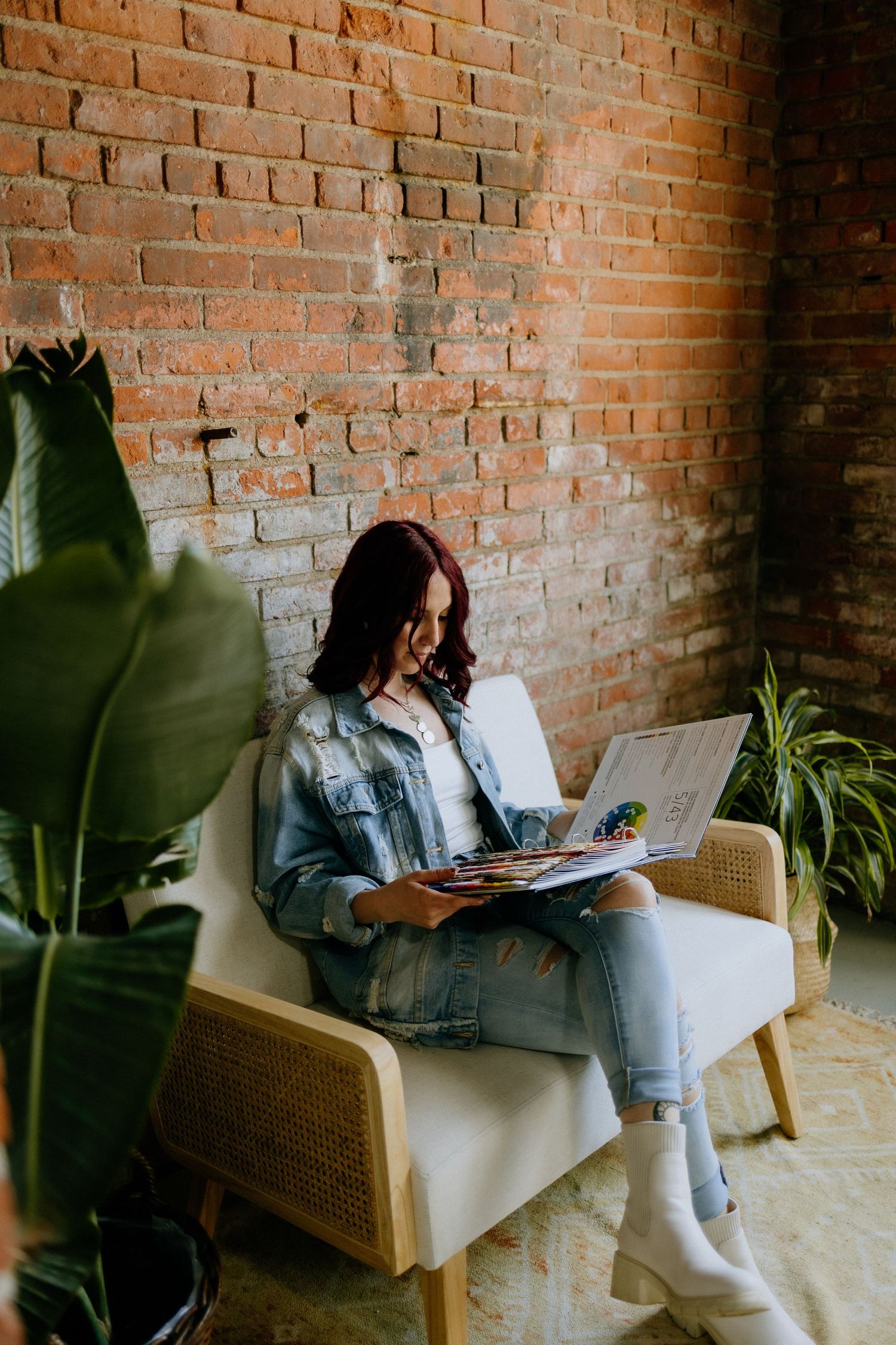 Person reading a book while relaxing in a modern chair against exposed brick wall with plants nearby.