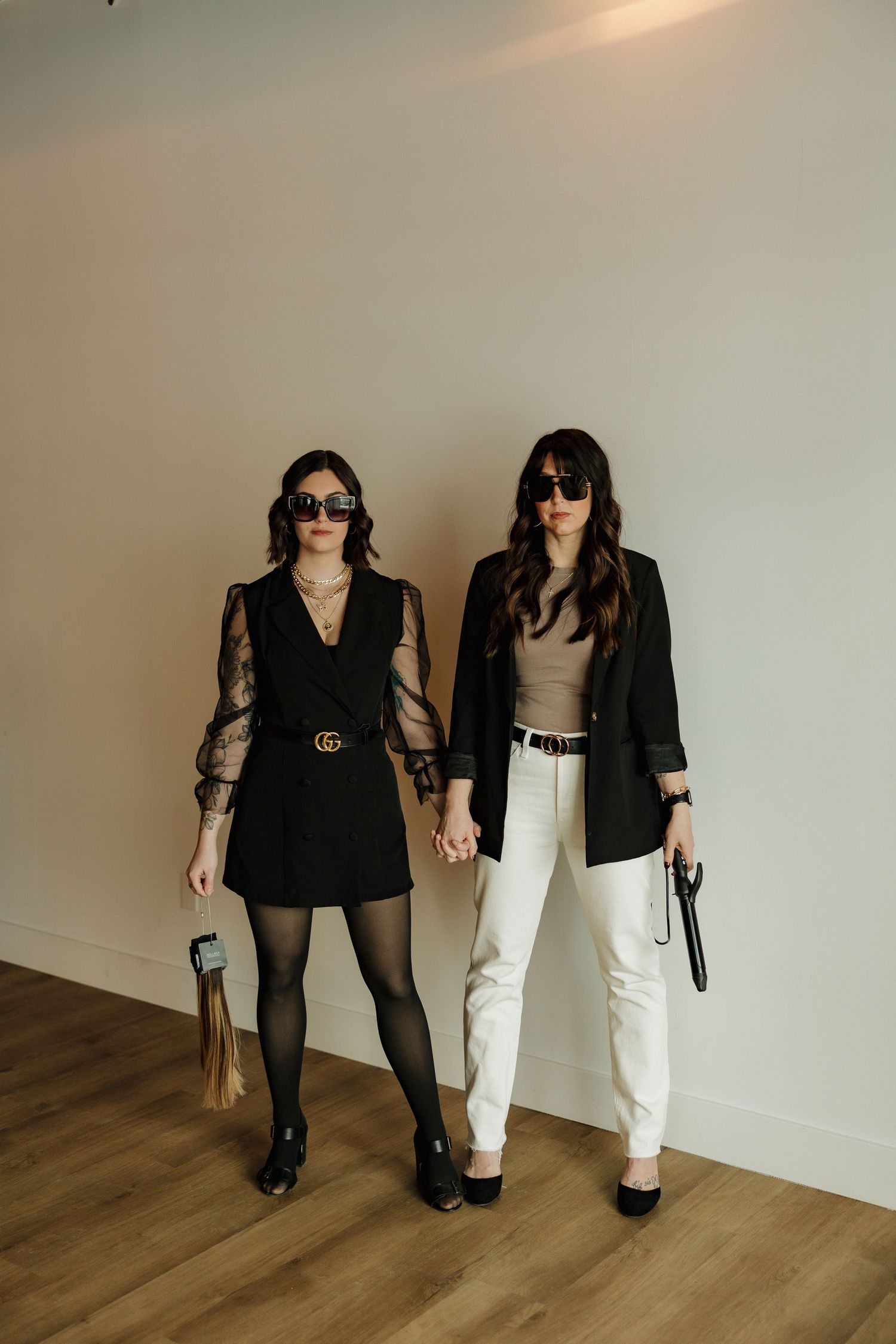 Two women pose in contrasting black and white outfits with coordinated blazers and sunglasses in a studio setting.