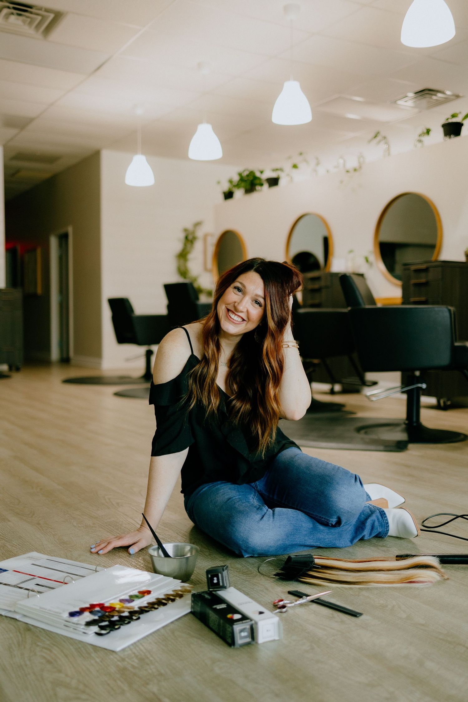 Person in black top sits on salon floor with styling products and materials.