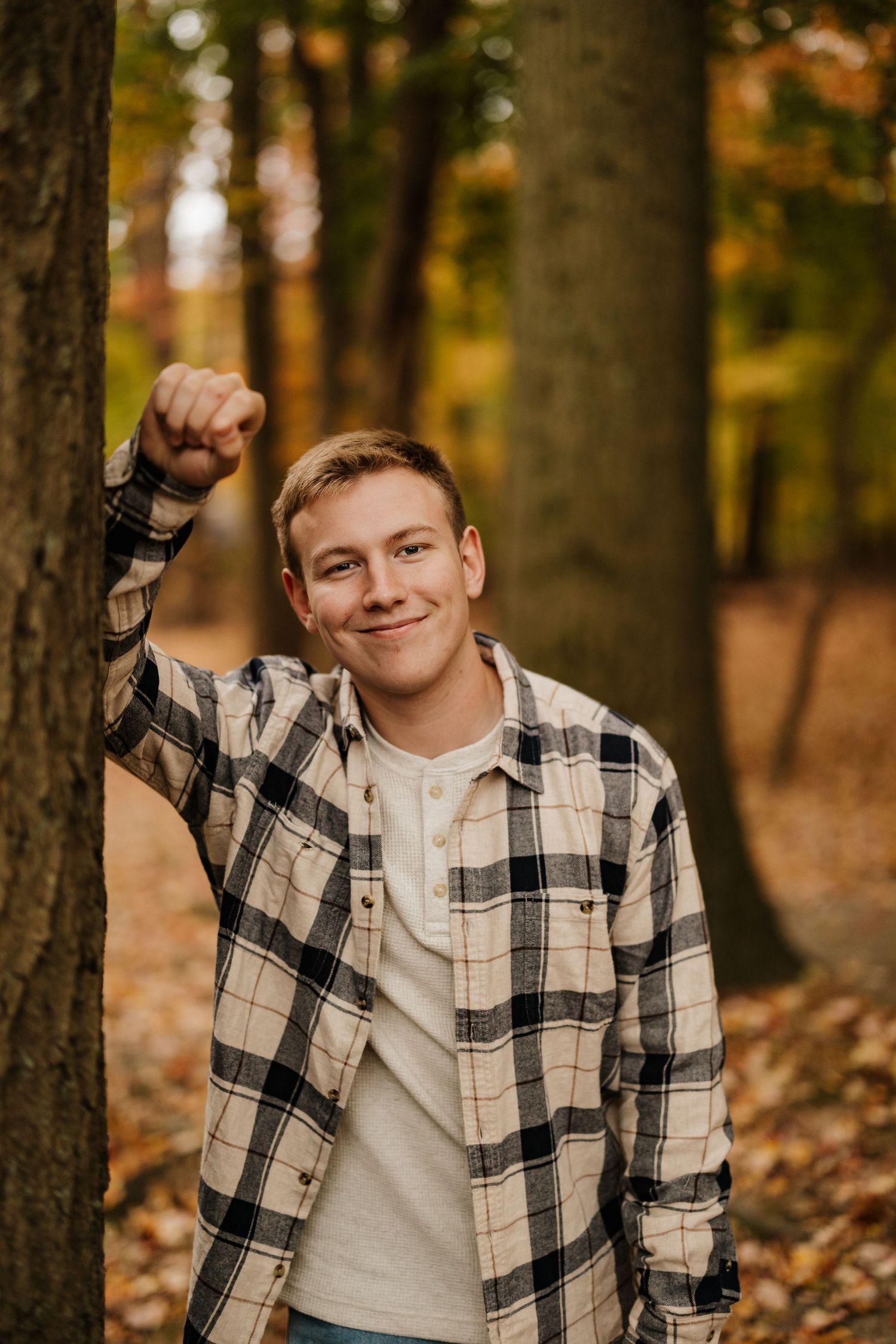Casual autumn portrait in plaid flannel shirt leaning against tree in forest setting.
