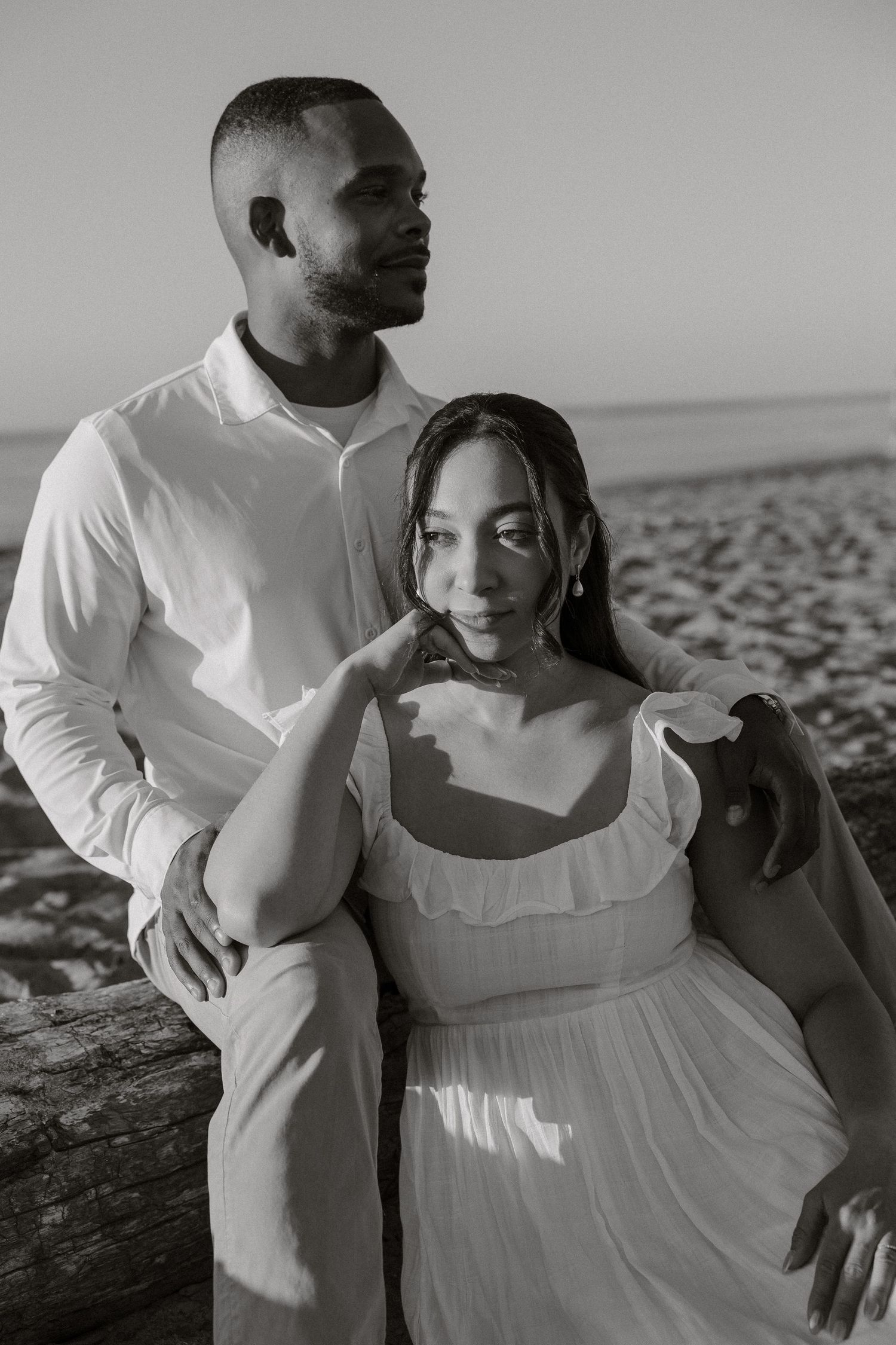 A romantic black and white beach portrait of a couple in white outfits embracing on the sand.
