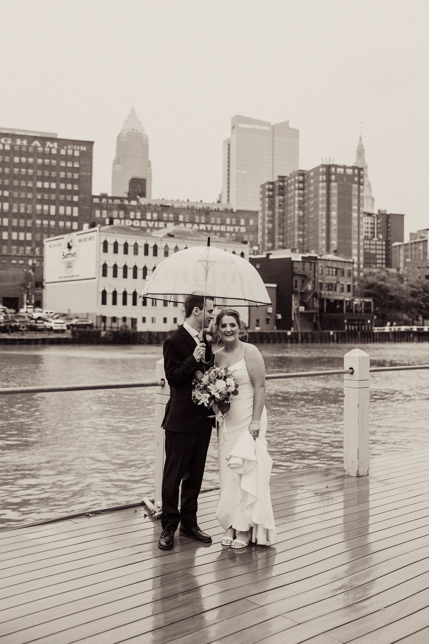 Wedding couple on a waterfront dock with city skyline in the background under cloudy skies.
