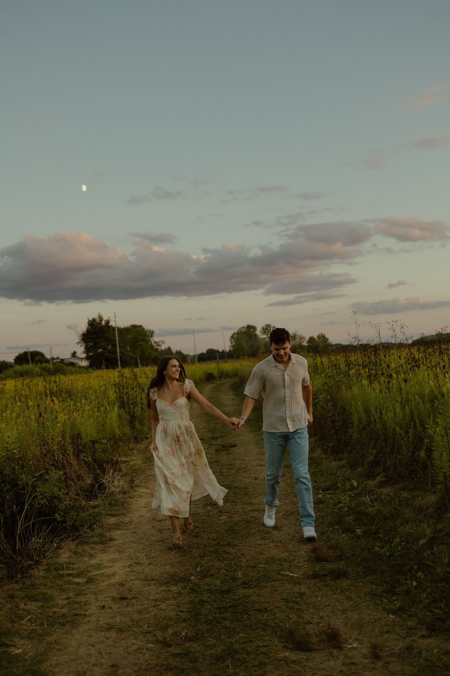Two people walk together through a golden field at sunset as clouds drift overhead in a romantic countryside scene.
