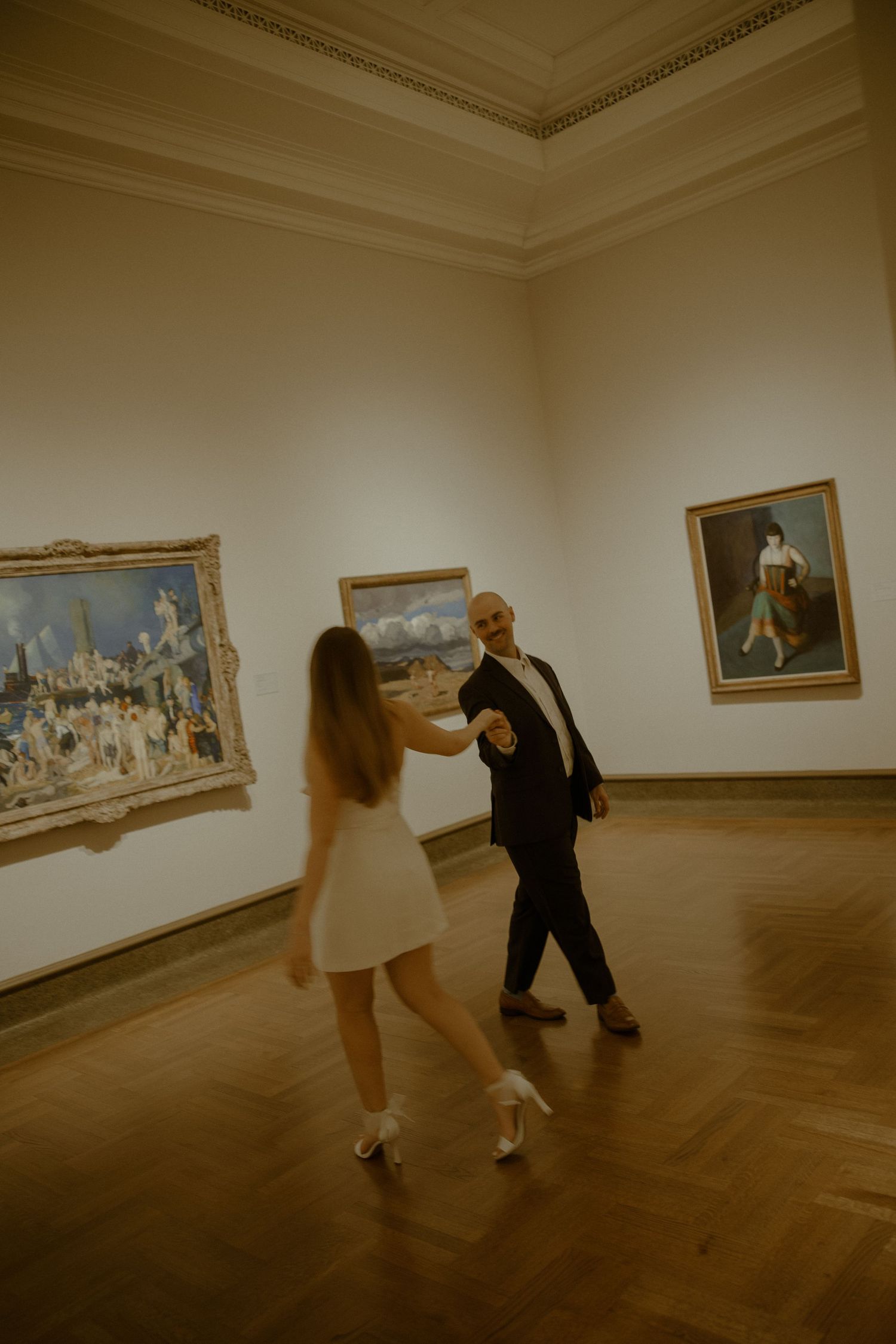 A couple dances together in an art museum gallery with classical paintings displayed on white walls.