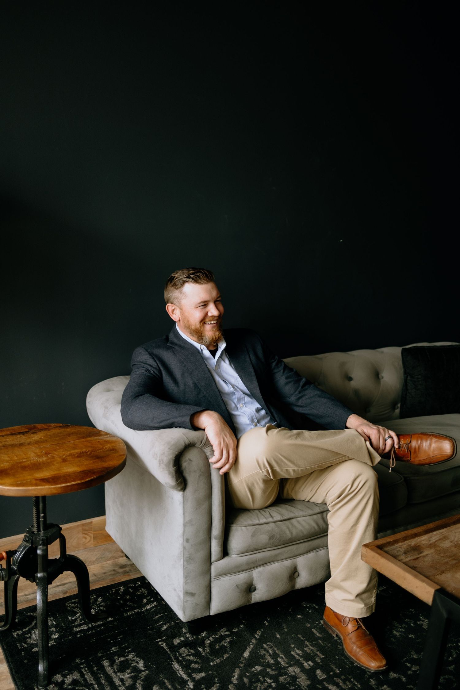 Business professional relaxing in gray armchair with wooden side table against dark background.
