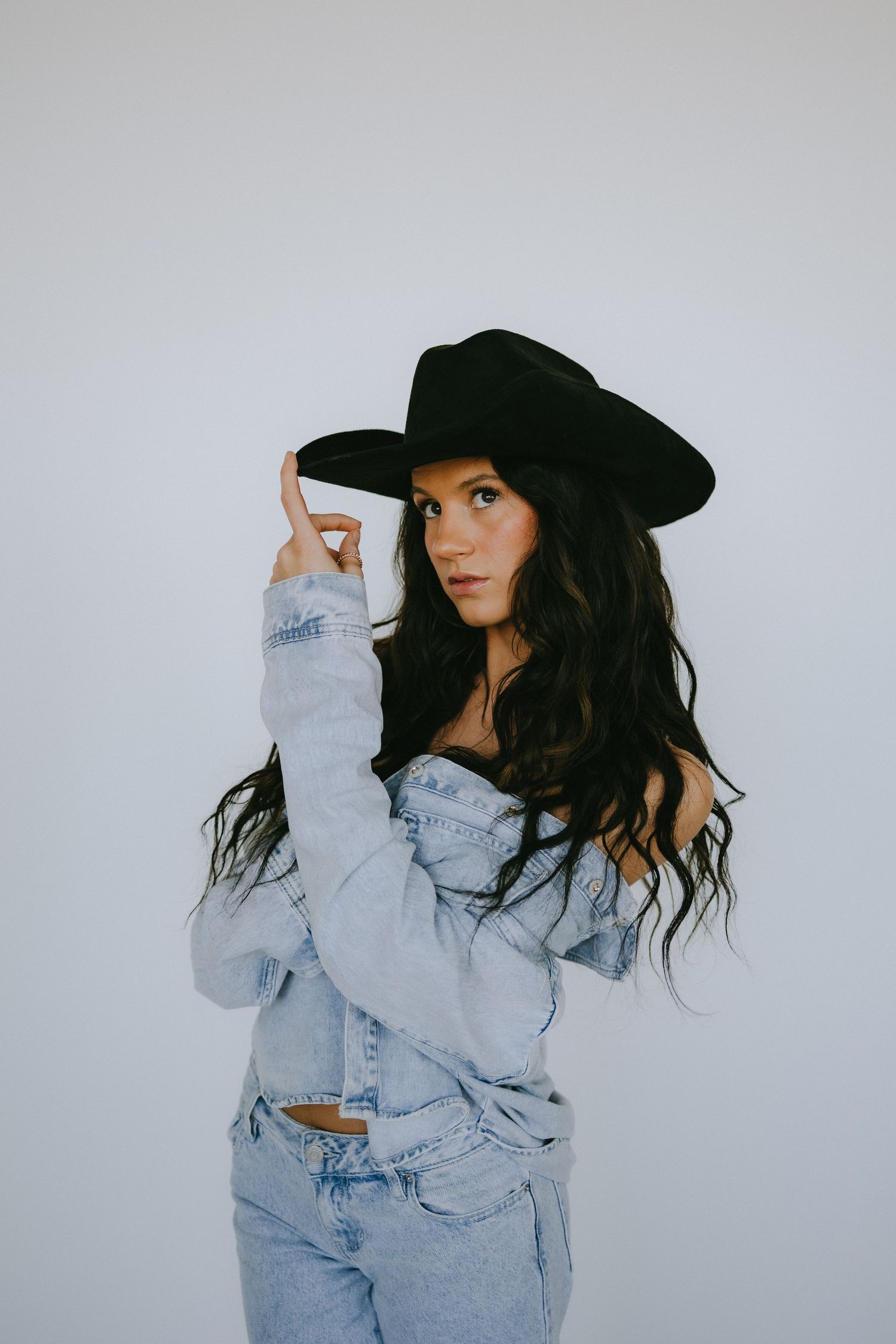 Stylish portrait with black cowboy hat and denim jacket against white studio background.