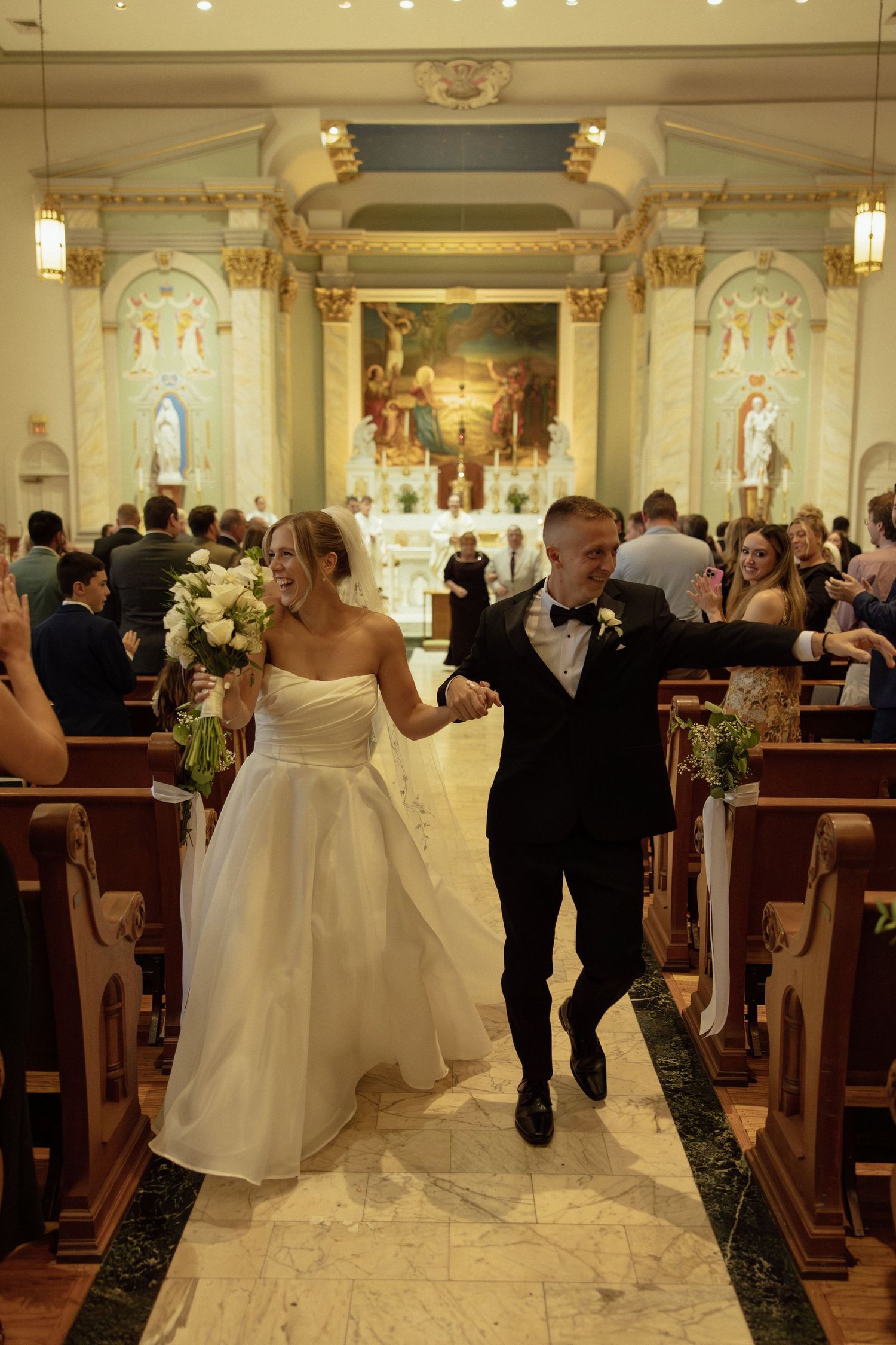 Wedding party walks down the aisle of an ornate church with elegant decor and floral arrangements.