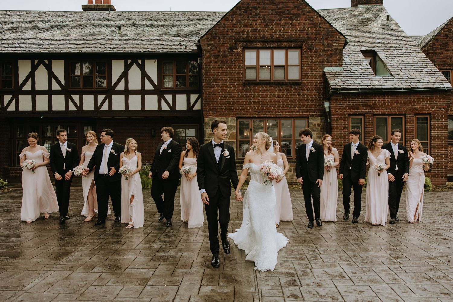 A wedding party poses in front of a Tudor-style brick building on a stone patio during an elegant outdoor celebration.