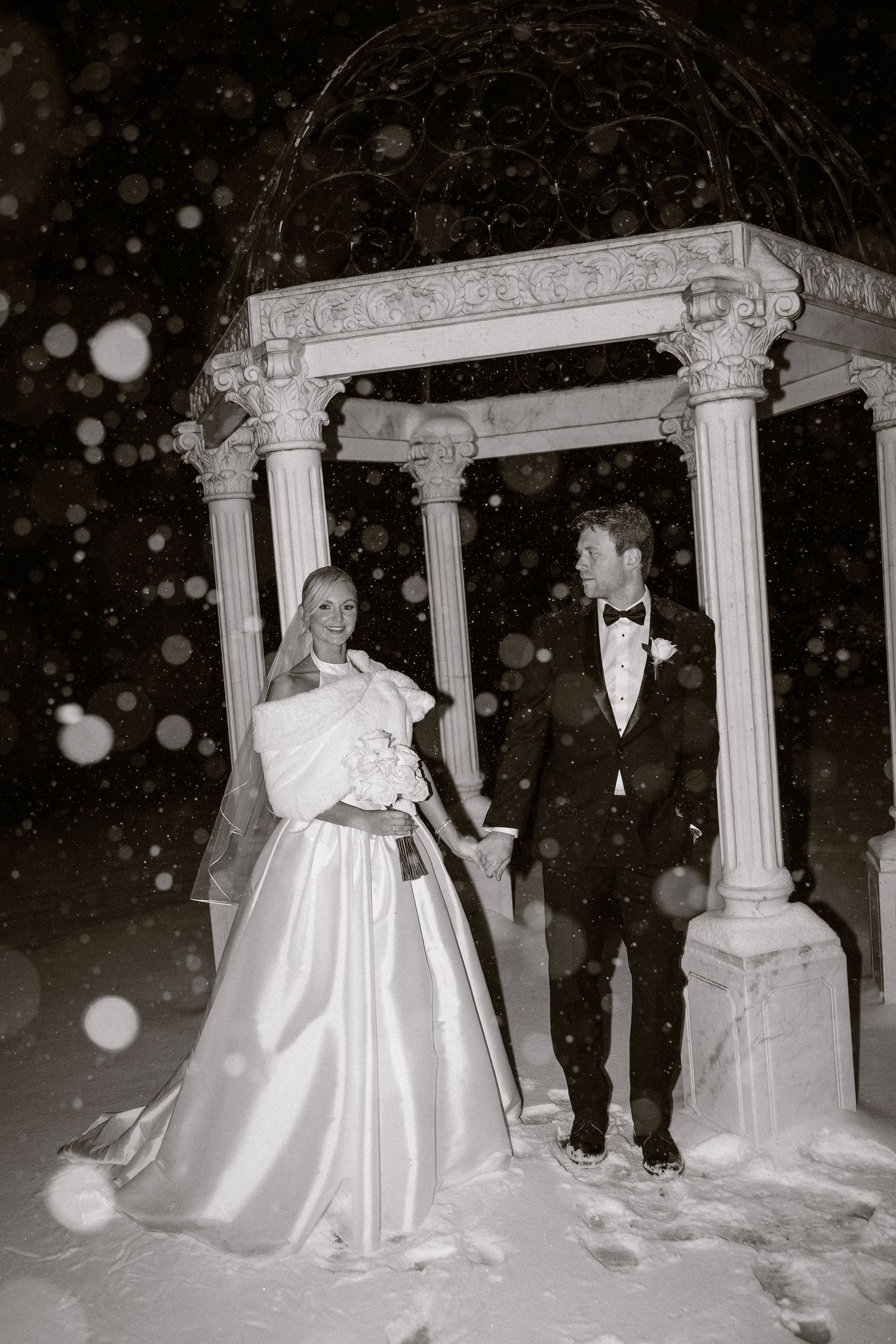 A bride in a white gown and groom in tuxedo standing between snow-covered columns.